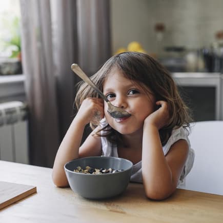 Fröhliches Kind sitzt mit Löffel im Mund vor der Müslischale.