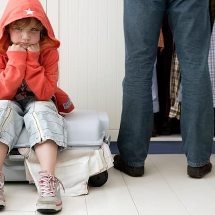 Schlecht gelauntes Kind sitzt auf Koffer, Mutter steht daneben.