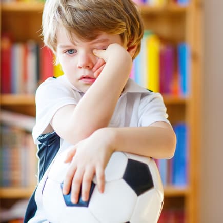 Krankes Kind reibt sich die Augen mit Fußball im Arm.