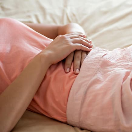 Frau liegt auf dem Bett und hält sich die Hände auf den Bauch.