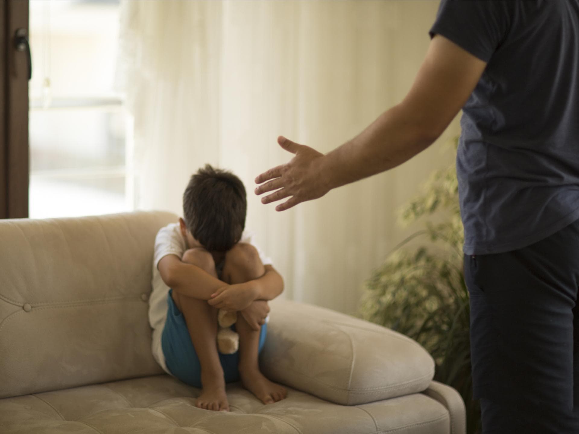 Junge sitzt mit angezogenen Knien auf dem Sofa, während sein Vater mit ihm schimpft.