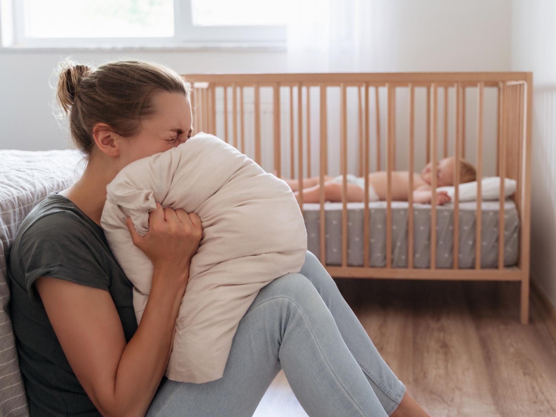 Eine Frau schreit in ein Kissen, während ihr Baby im Bett schläft