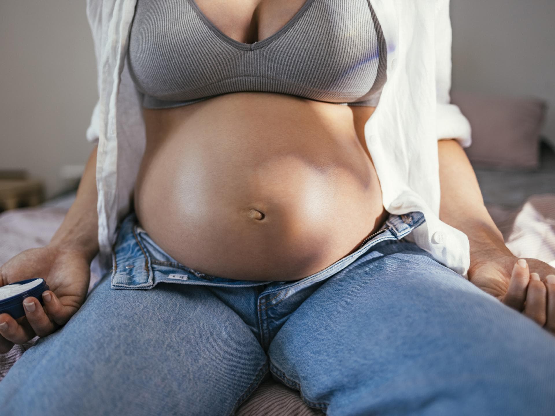 Eine Frau mit Babybauch sitzt mit geöffneter Hose auf dem Bett