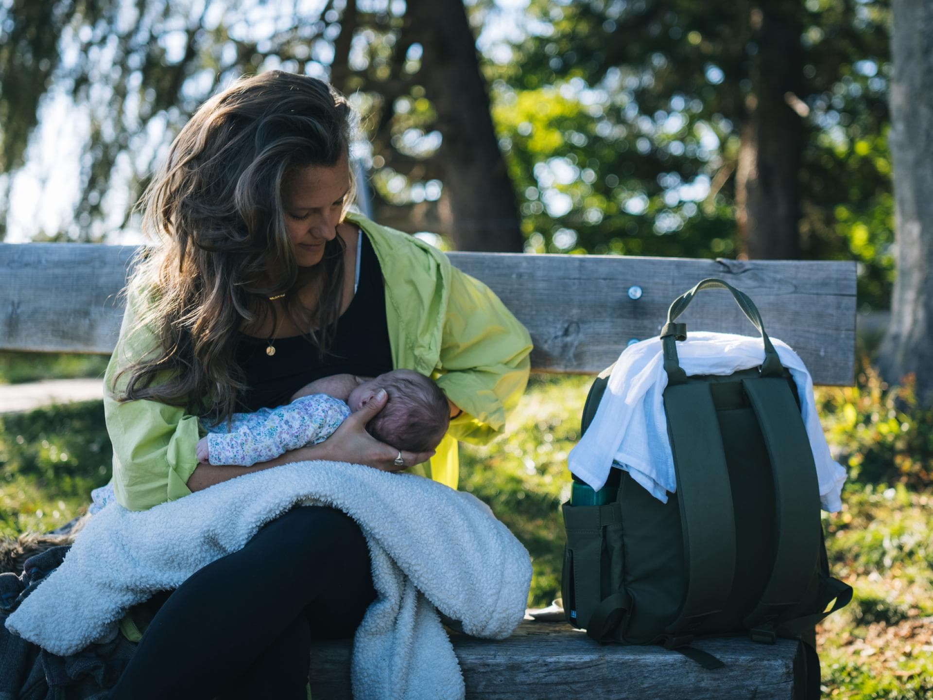 Wickelrucksack packen: Das gehört wirklich rein! Eine Frau, die ihr Baby auf einer Bank im Grünen stillt.