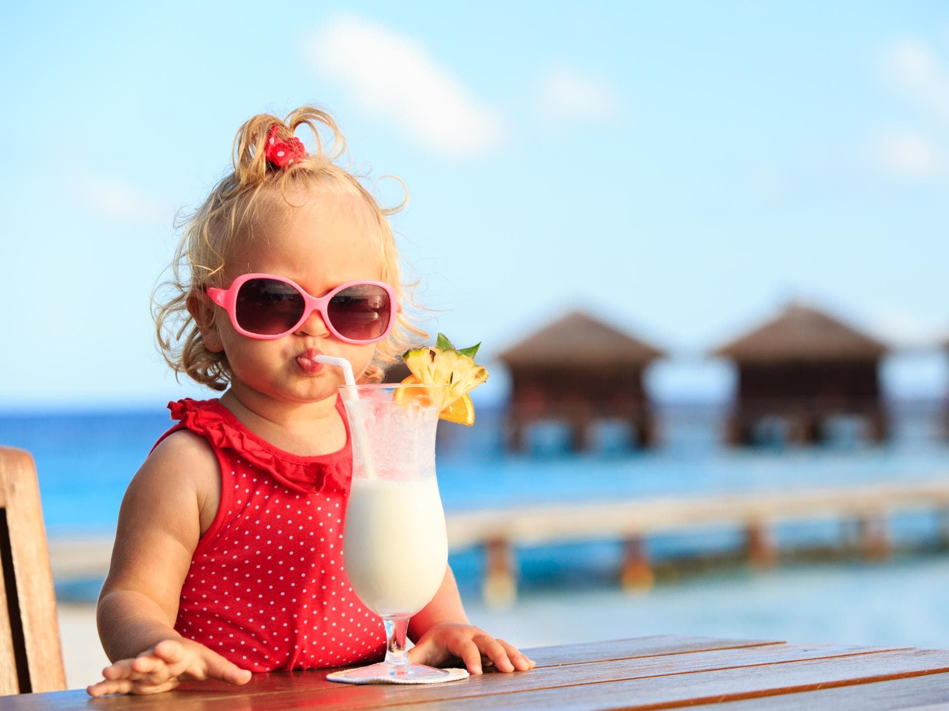 Juni-Babys sind echte Sonnenkinder. Ein kleines Mädchen mit rotem Badeanzug und Sonnenbrille trinkt einen Kinder-Cocktail.