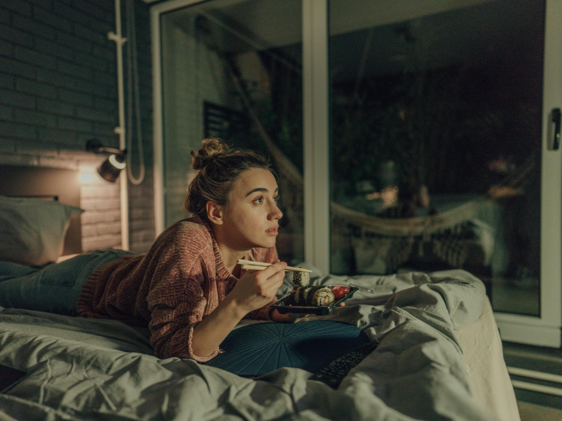Eine Frau liegt abends im Bett, schaut TV und isst Sushi