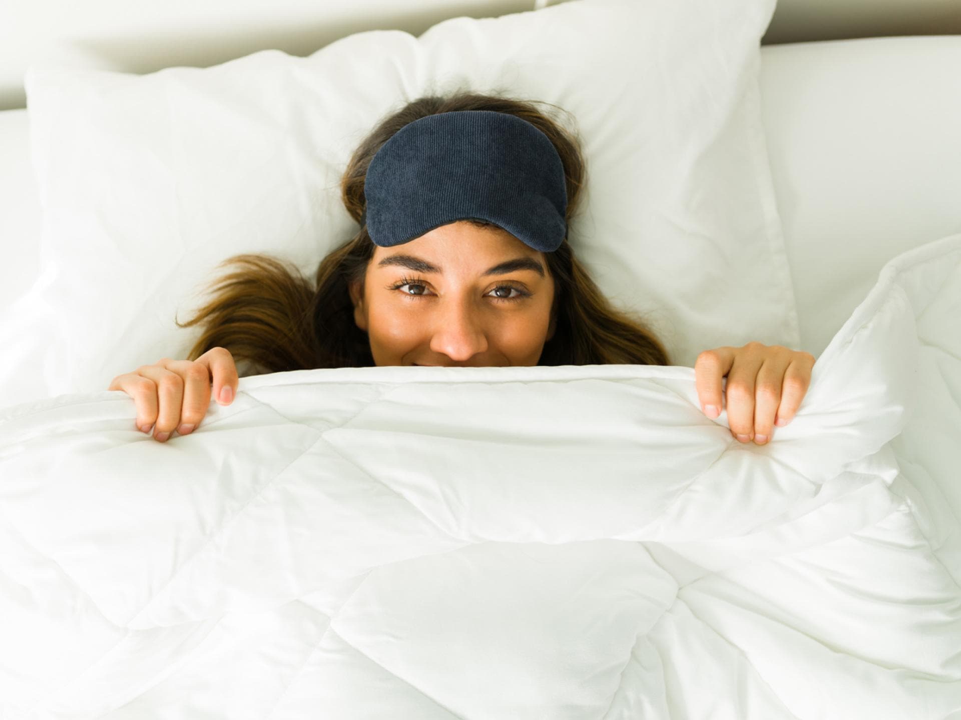Eine Frau liegt mit Schlafbrille im Bett und zieht sich die Decke übers Gesicht.