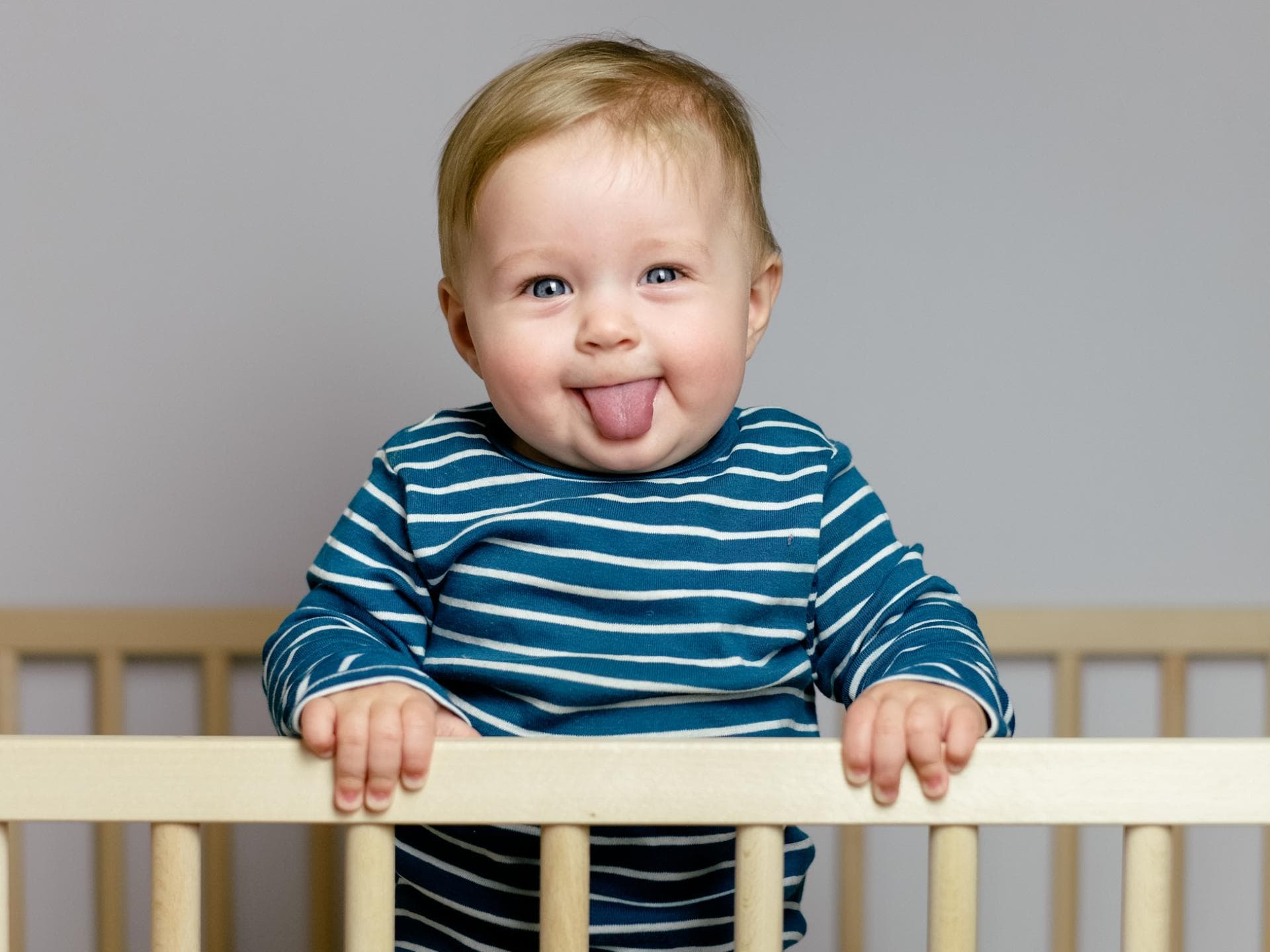 Ein kleiner Junge steht in seinem Gitterbett und streckt die Zunge raus.