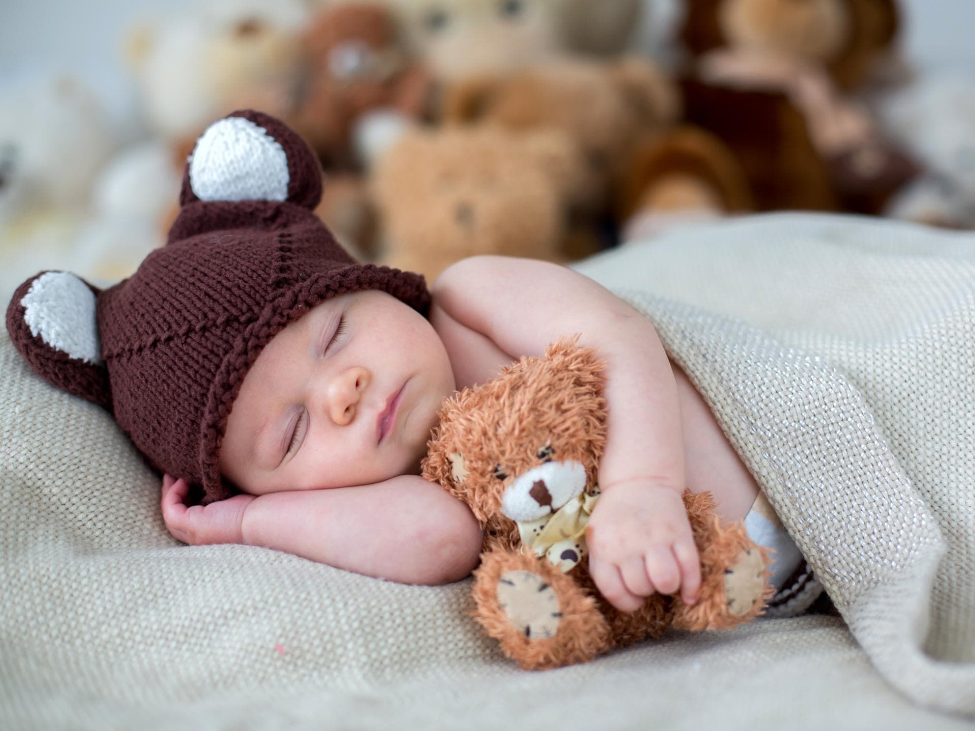 Ein Baby liegt eingekuschelt auf dem Bett und schläft mit Teddybär im Arm.
