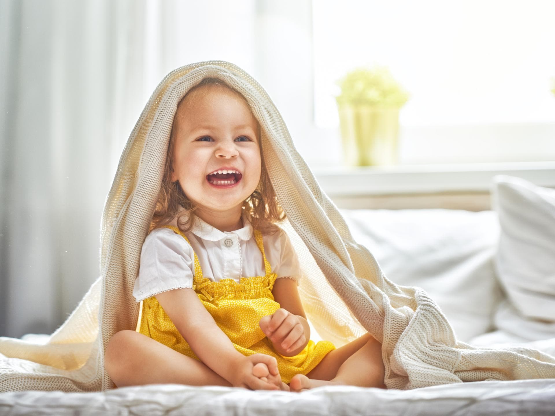 Ida oder Ivy? Mädchennamen mit I klingen häufig verspielt. Ein kleines Mädchen sitzt lachend auf dem Bett und hat sich die Bettdecke über den Kopf geworfen.