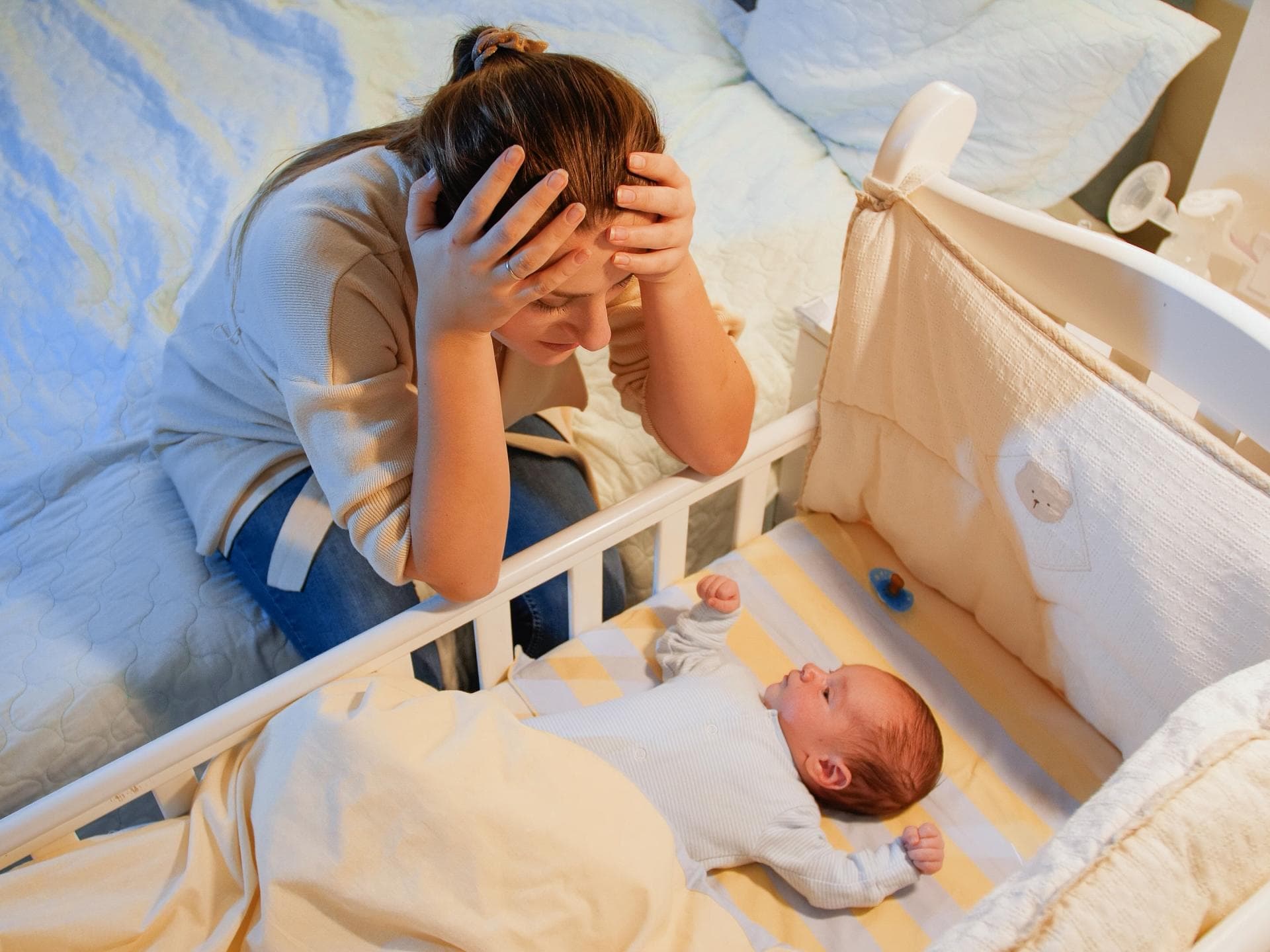 Mutter sitzt am Babybett und stützt ihren Kopf in ihre Hände.