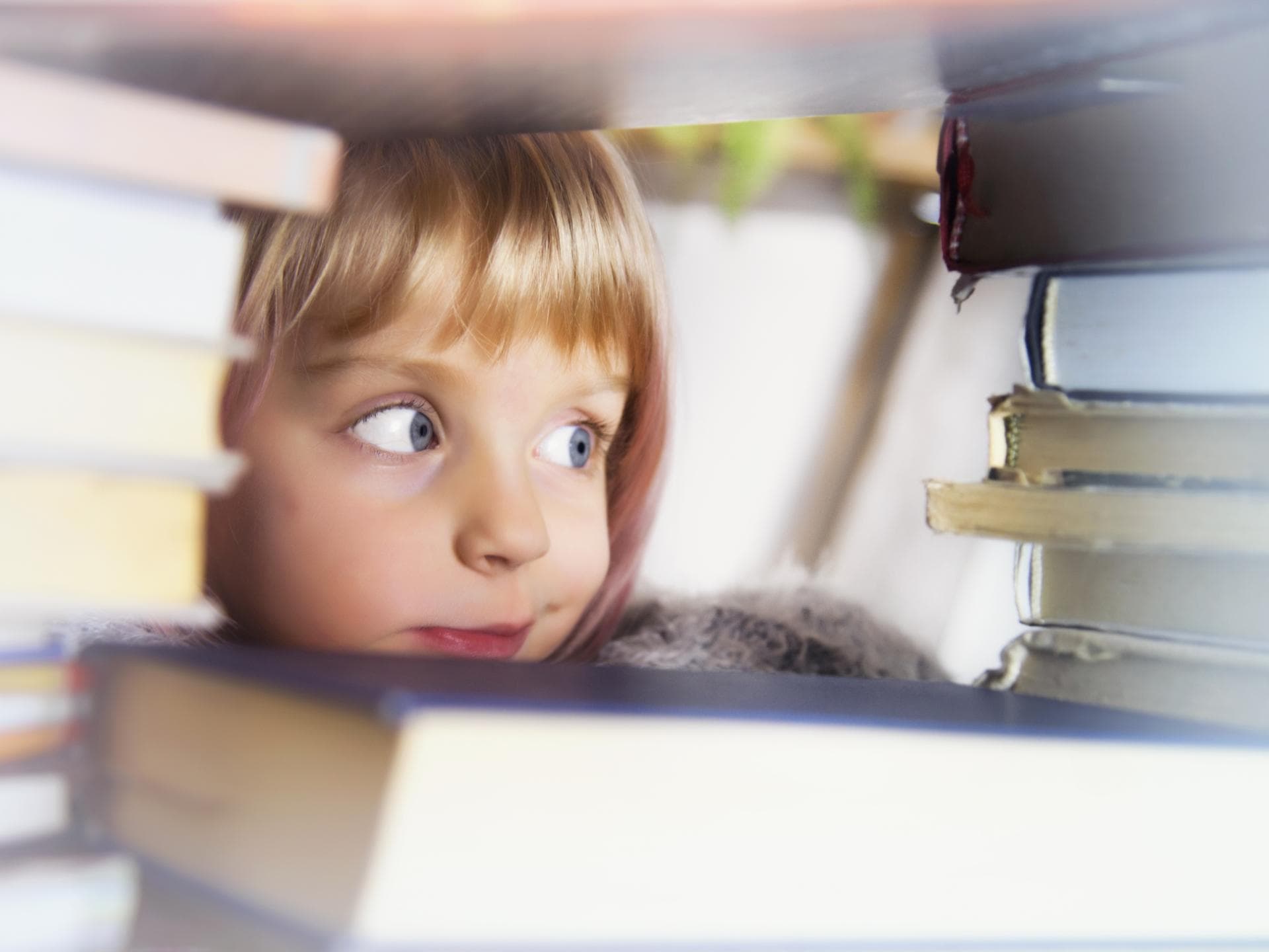 Mädchen schaut mit großen Augen zwischen Bücherstapeln hindurch