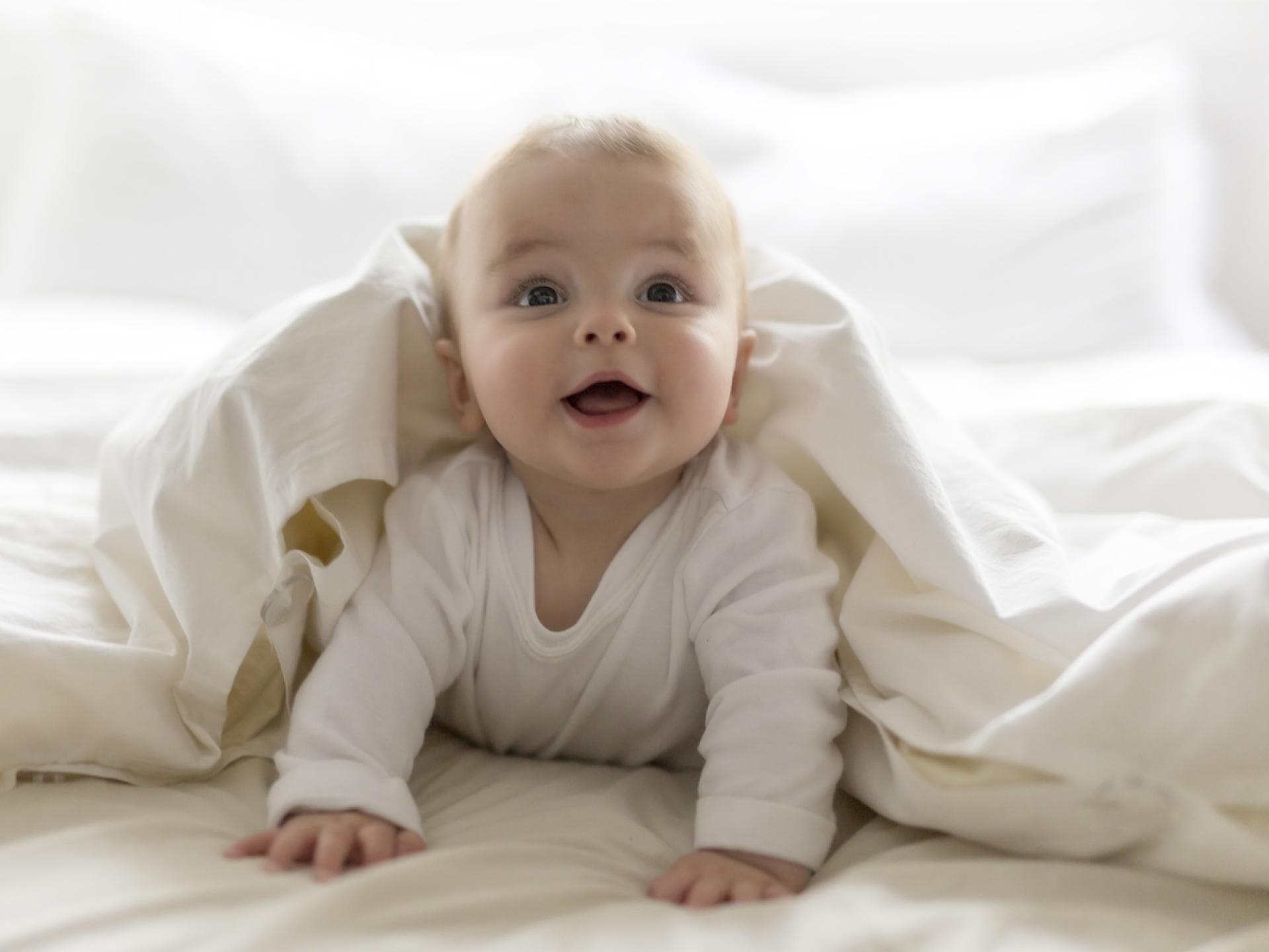 Ein kleiner Baby-Junge guckt frech unter einer weißen Bettdecke hervor. 