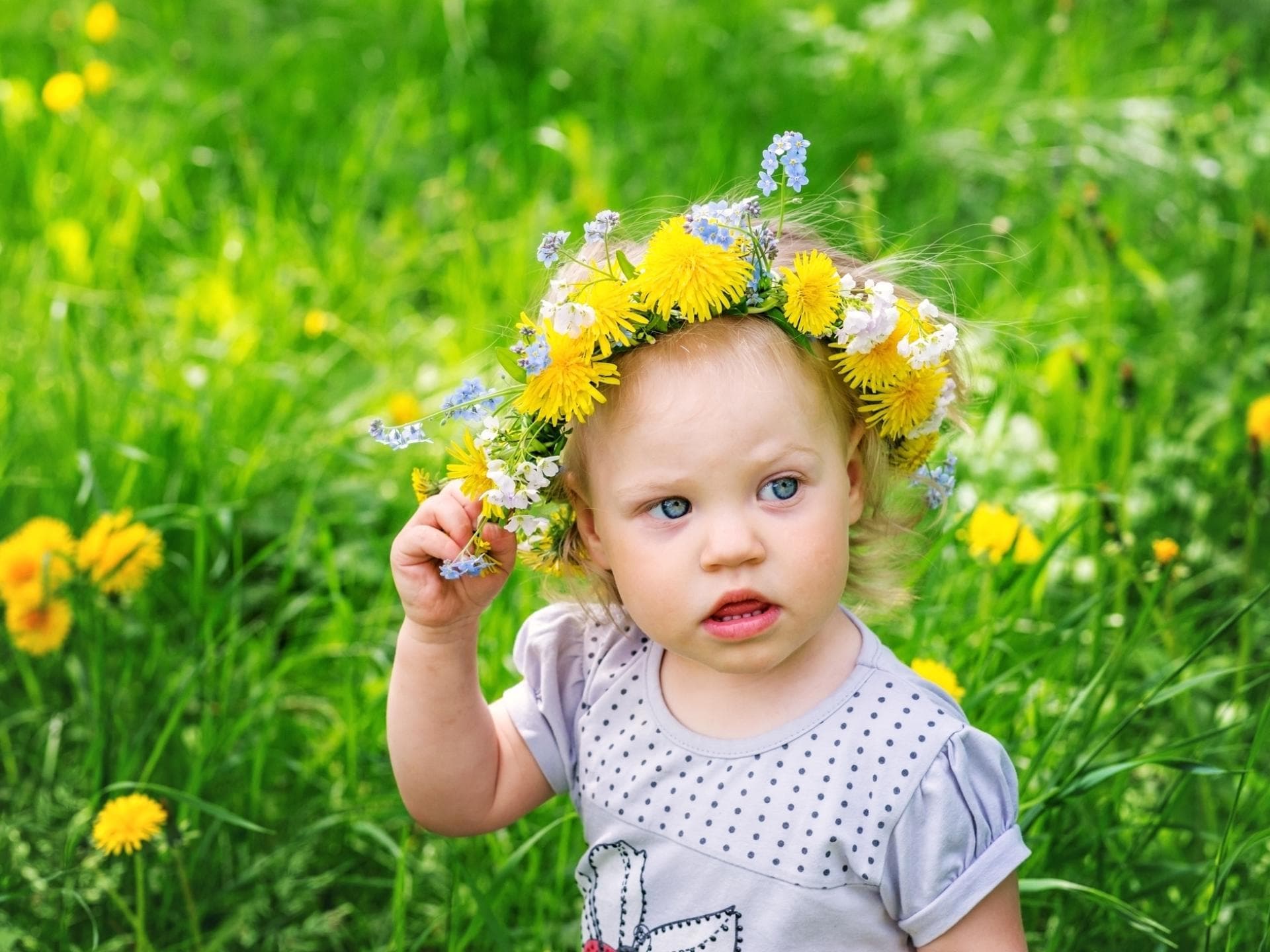 Kleines Mädchen mit Blumenkranz auf dem Haar steht auf einer Wiese.