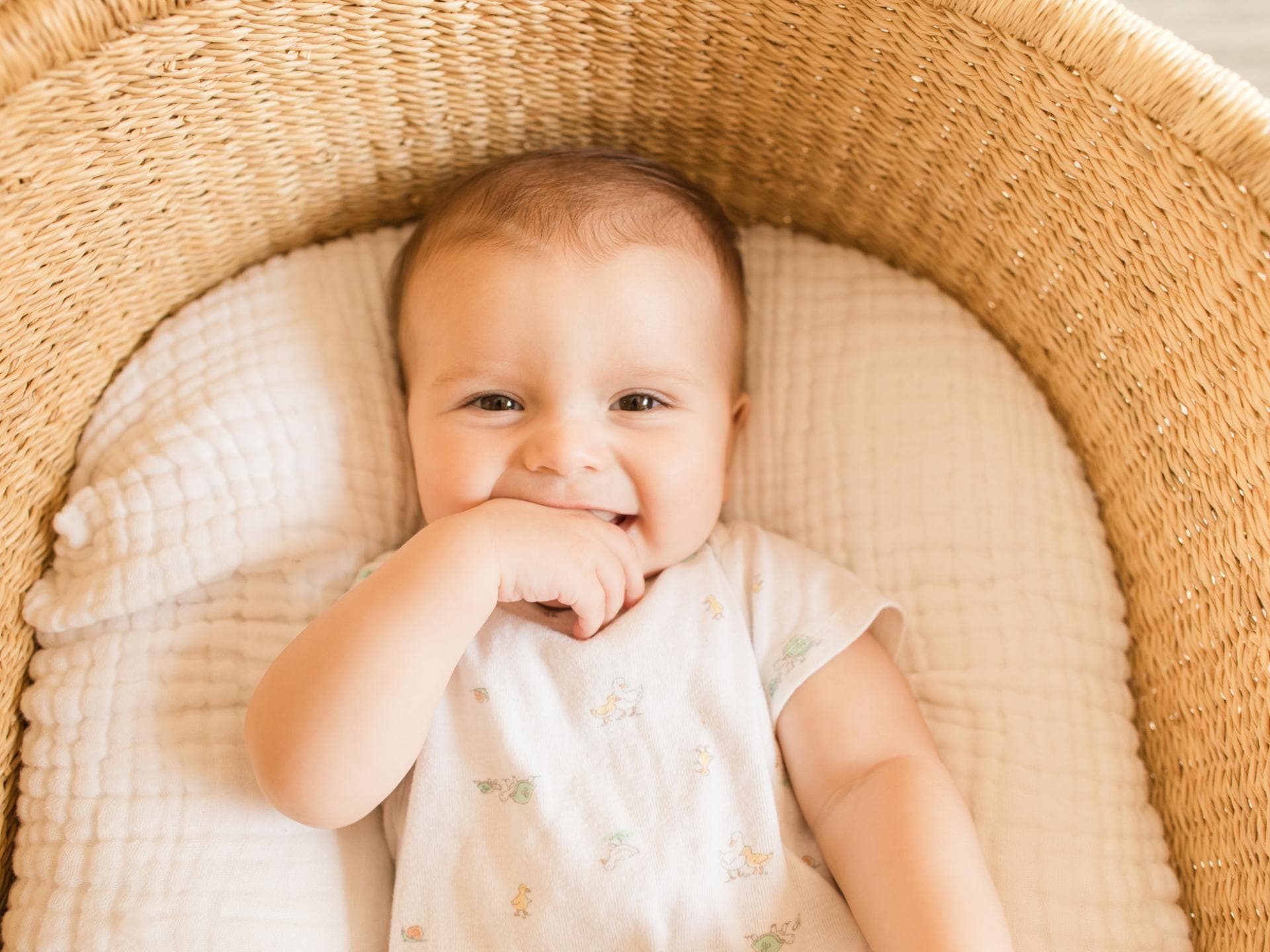 Ein kleiner, süßer Junge, der seine Finger im Mund hat und vom Babybett in die Kamera grinst.