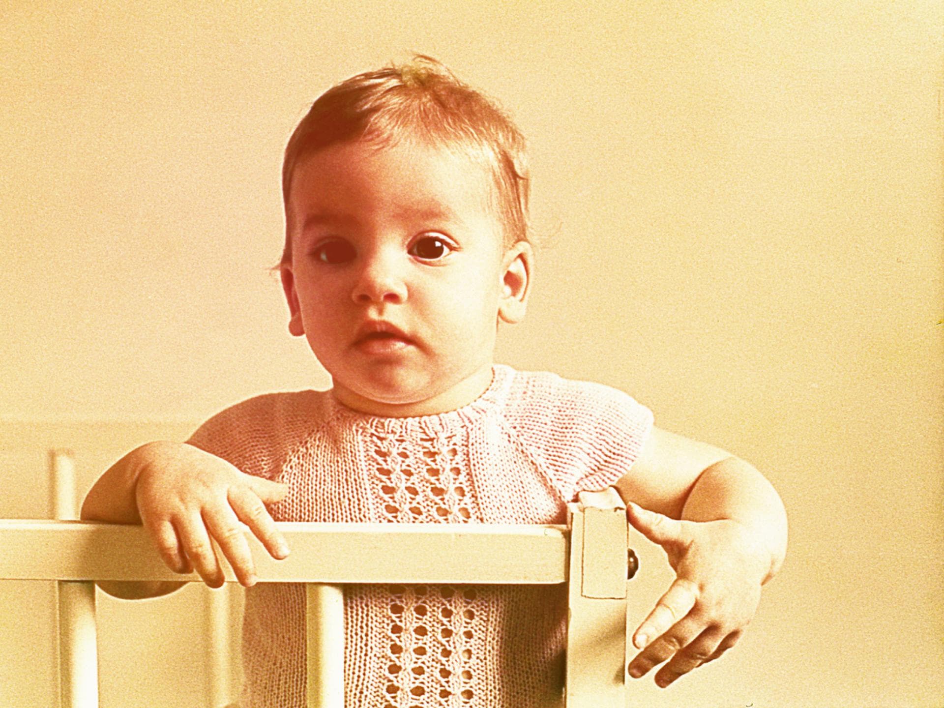 Ein Baby-Mädchen in den 60ern steht niedlich im Gitterbett