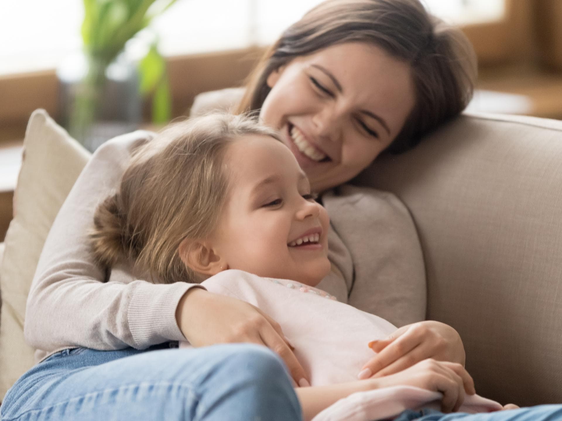 Mama kuschelt und lacht mit Tochter auf Sofa.