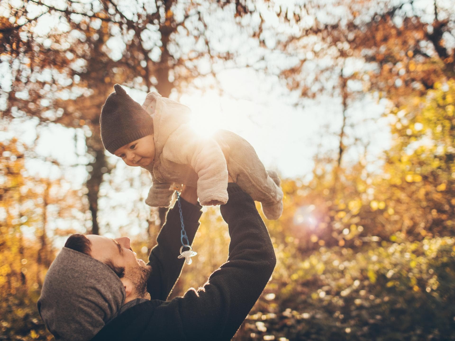 Was ist die richtige Babykleidung im Herbst? Papa hält Baby hoch im herbstlichen Wald