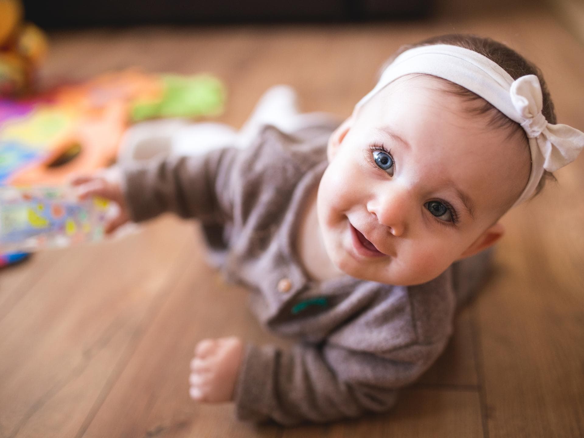 Kleines Baby-Mädchen mit Haarband liegt auf dem Boden und grinst.