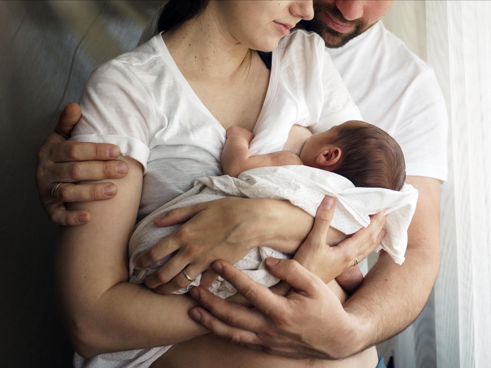 Frau hält Baby auf dem Arm und stillt es, Mann steht hinter ihr und umarmt beide. Wie schmeckt eigentlich Muttermilch? Das fragen sich manche Väter.