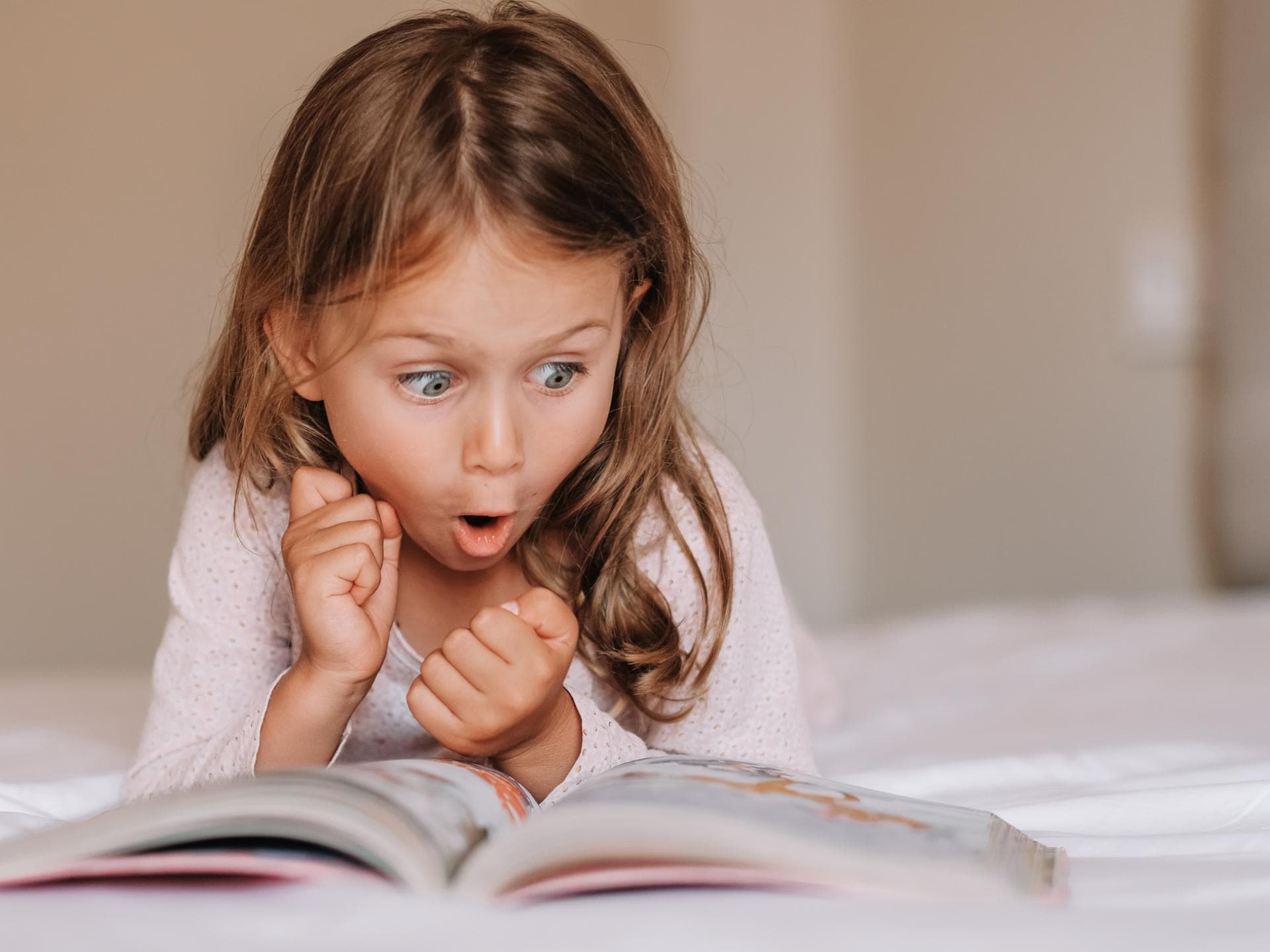 Kleines Mädchen liegt auf dem Bauch und schaut staunend ein Buch an. Je spannender ein Buch, desto eher wollen Erstleser weiterlesen!