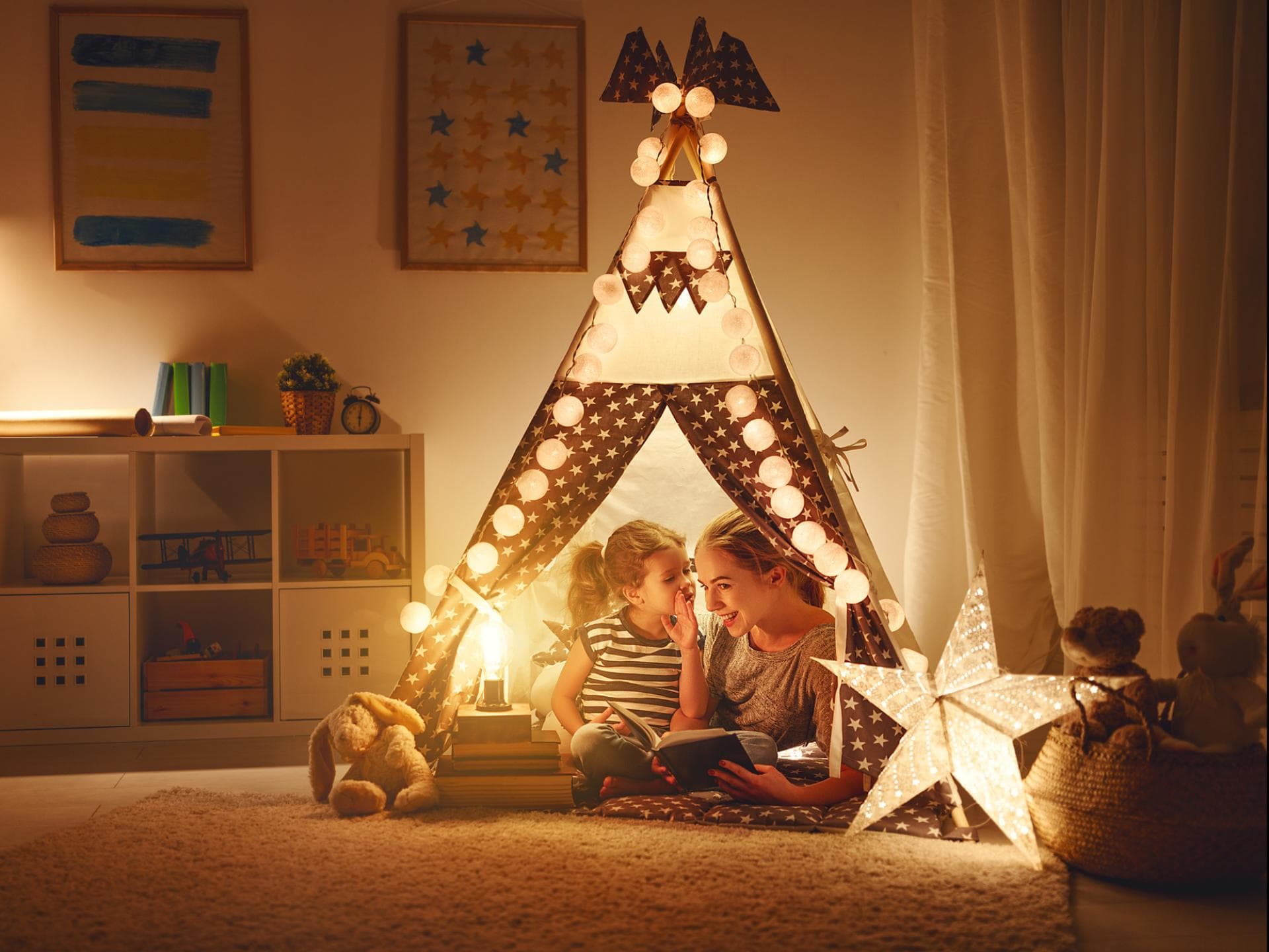 Tochter und Mutter sitzen im Tipi im aufgeräumten Kinderzimmer bei gemütlichem Licht.
