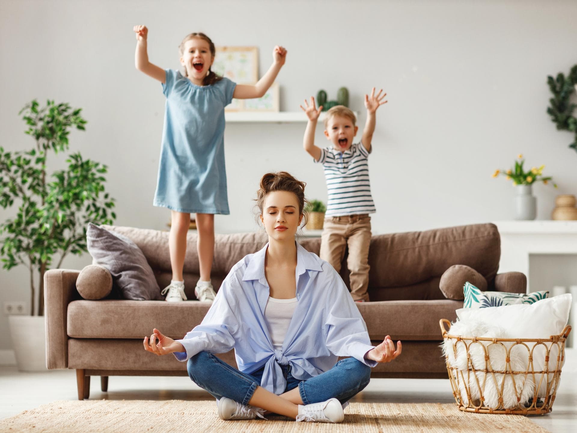 Mehr Entspannung im Alltag wünschen sich die meisten Eltern. Kinder springen auf dem Sofa, Mutter meditiert.