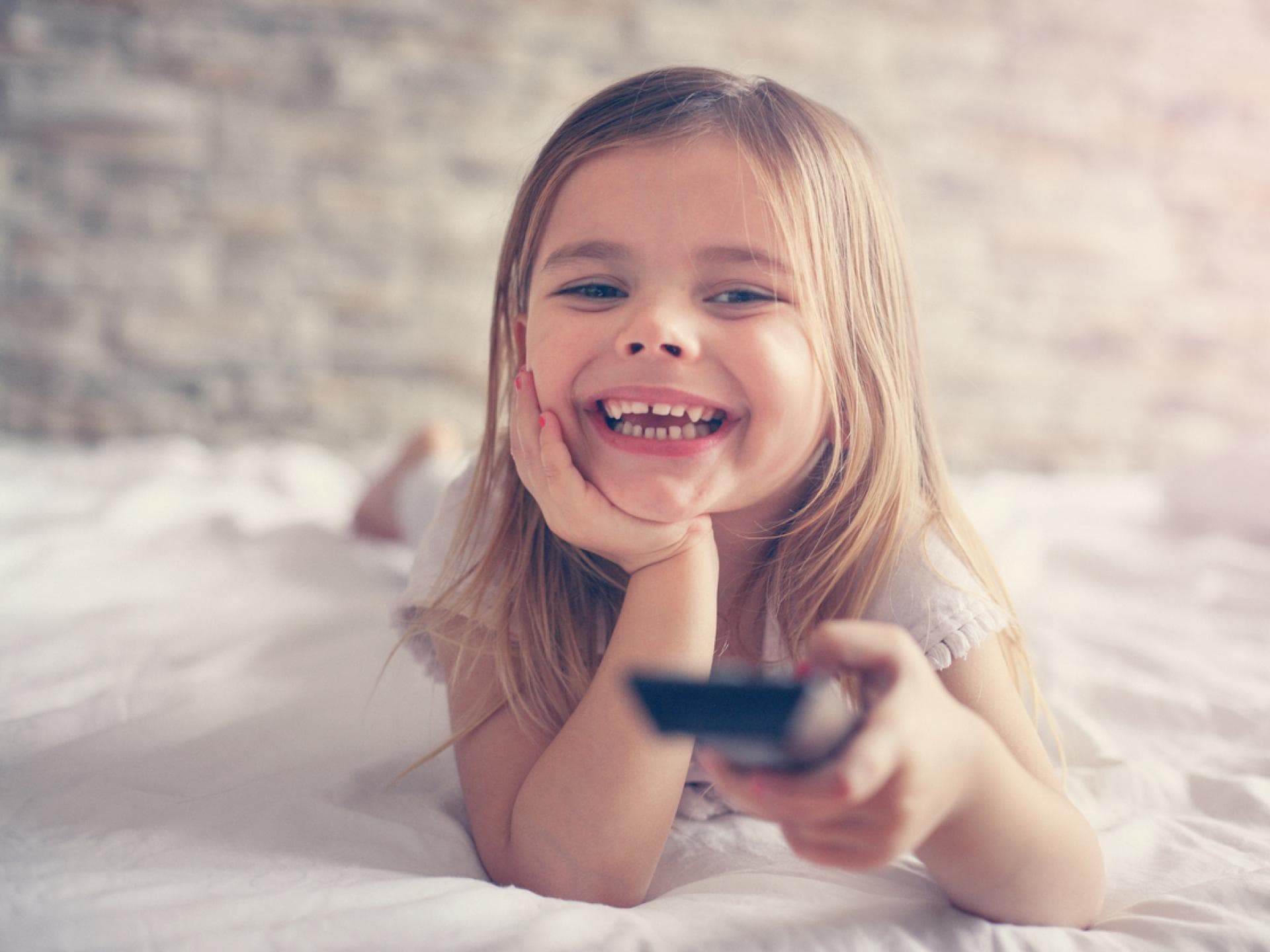 Medienzeit für Kinder stellt Eltern oft vor große Herausforderungen. Lachendes Mädchen liegt mit Fernbedienung im Bett.