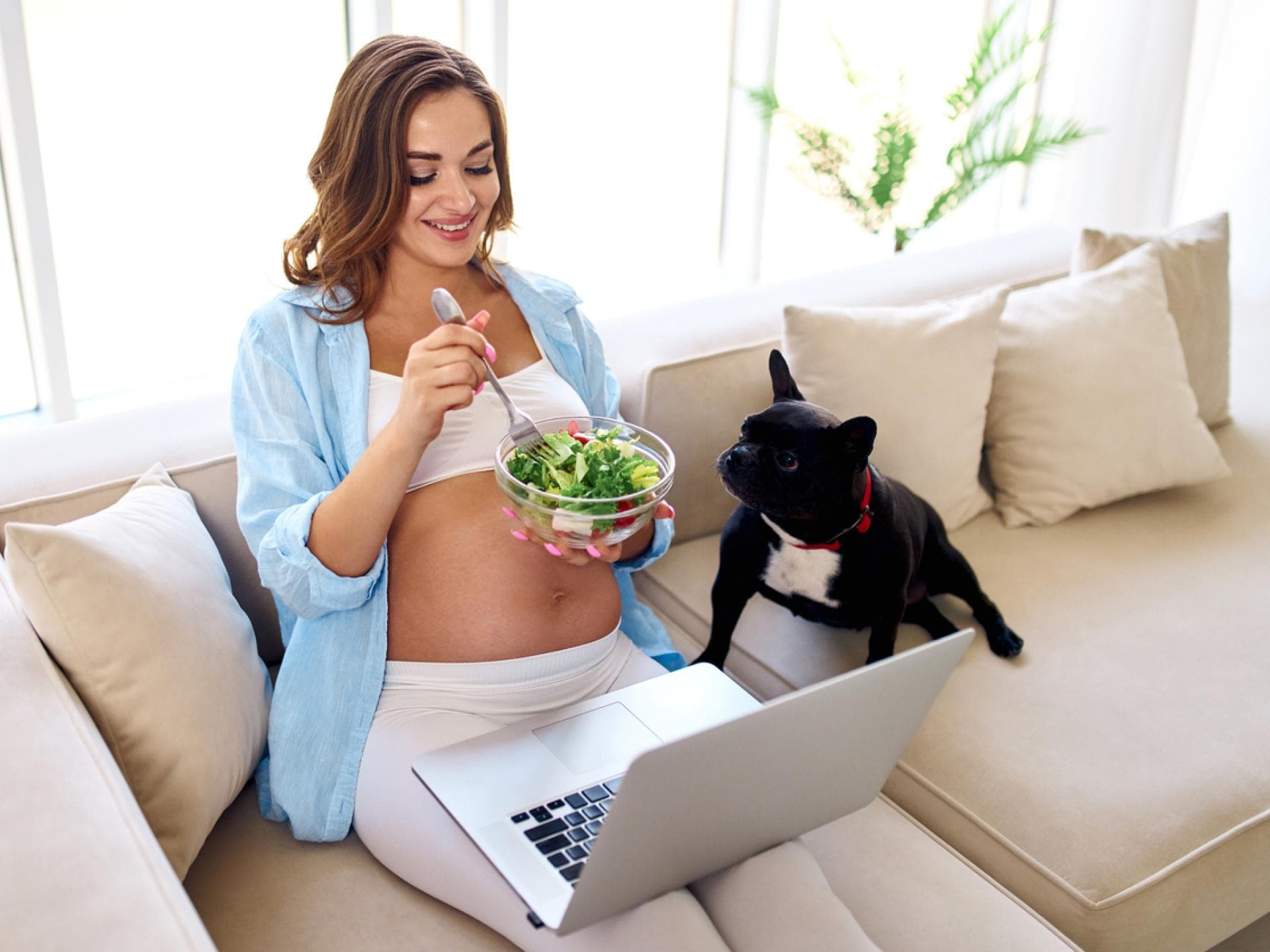 Schwangere sitzt auf Sofa und isst Salat.