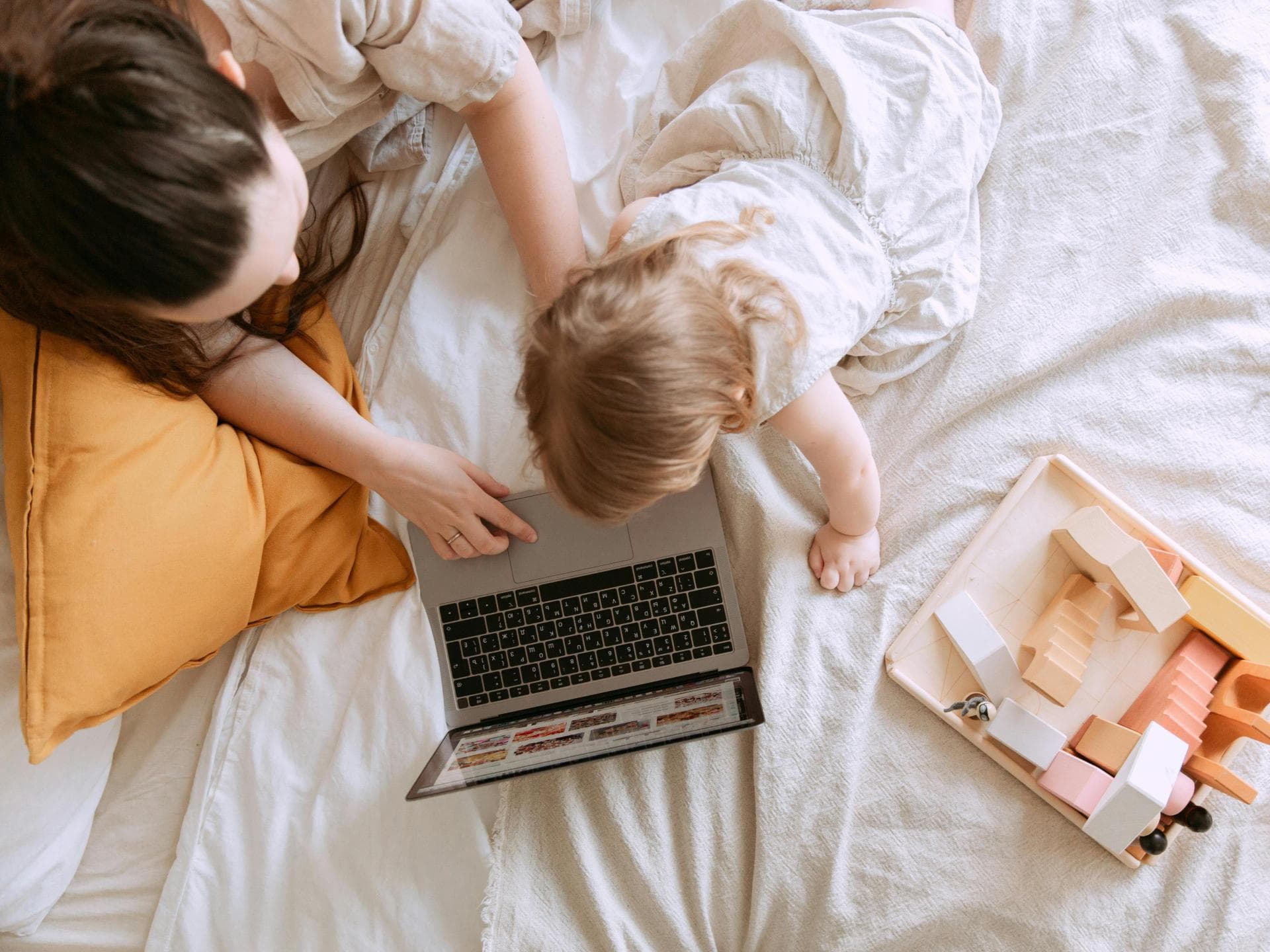 Frau und Kleinkind sitzen auf dem Bett und schauen auf einen Laptop, daneben ein Spiel-Tablett.