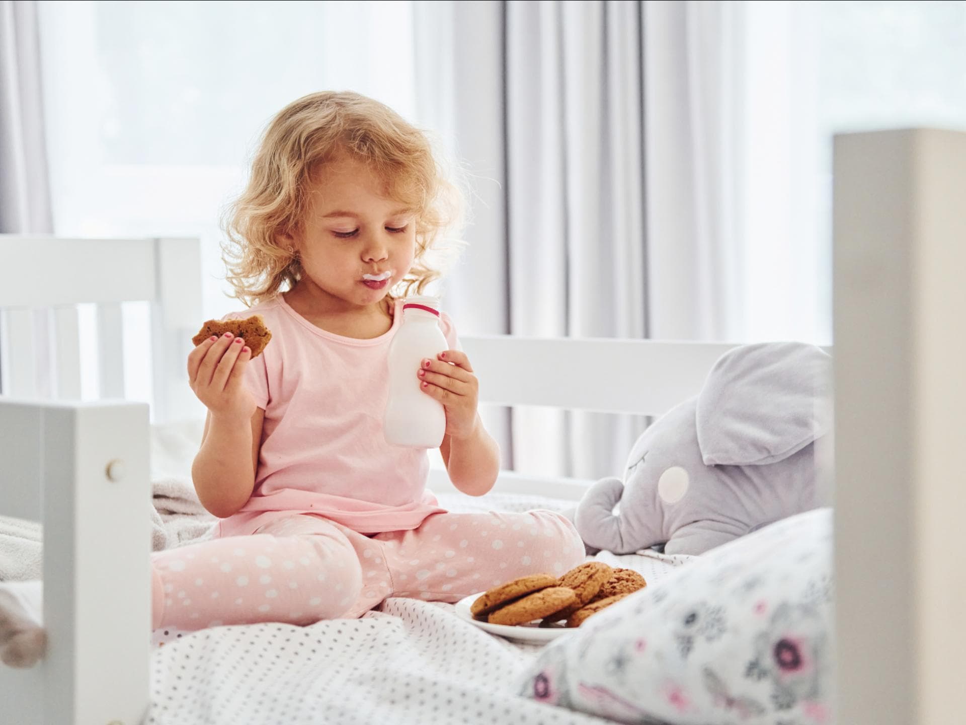 Ein kleines Mädchen isst im Bett Kekse und trinkt Milch.
