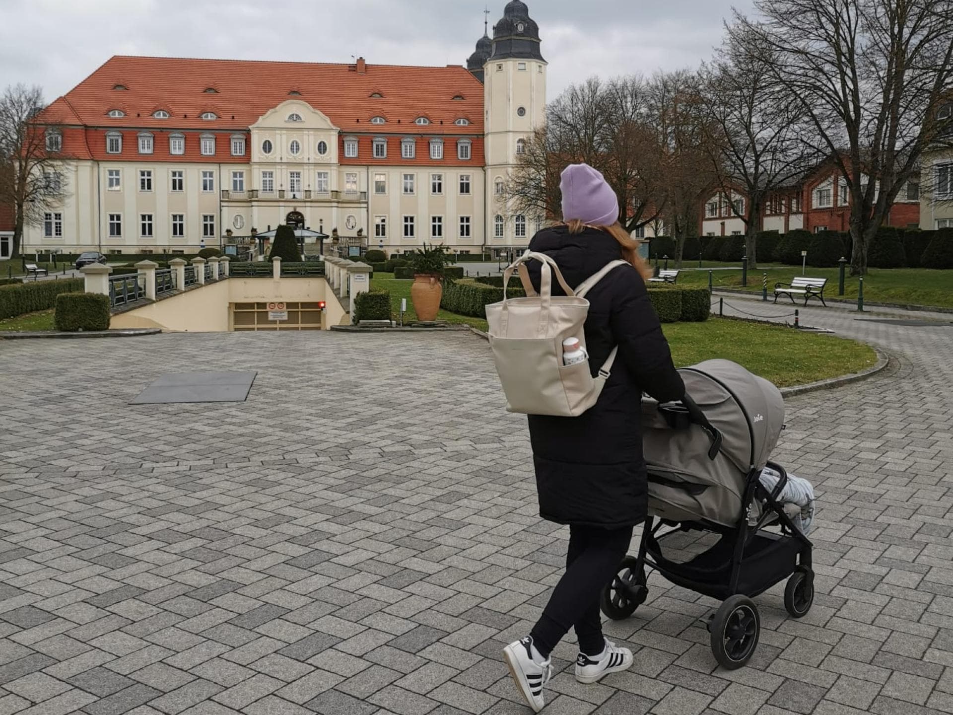 Wickelrucksäcke sind für Eltern unabdingbar. Eine junge Mutter läuft mit ihrem Kapten & Son-Wickelrucksack und Buggy vor einem Schloss.
