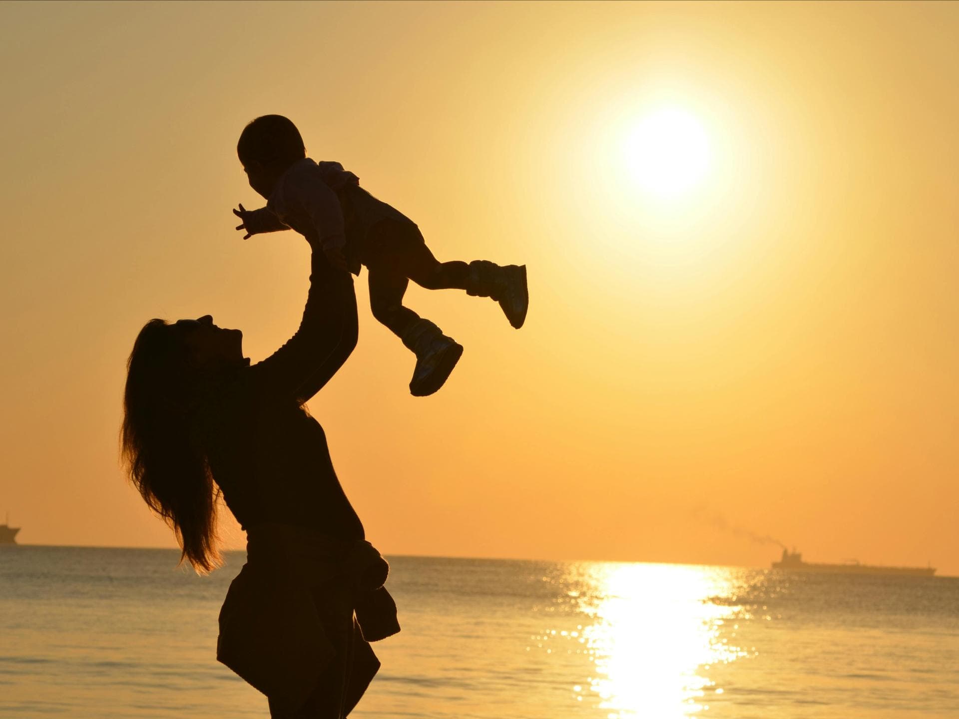 Jungennamen mit der Bedeutung Sonne sind bei Eltern sehr beliebt. Eine Mutter hält ihr Baby beim Sonnenuntergang am Strand in die Luft.