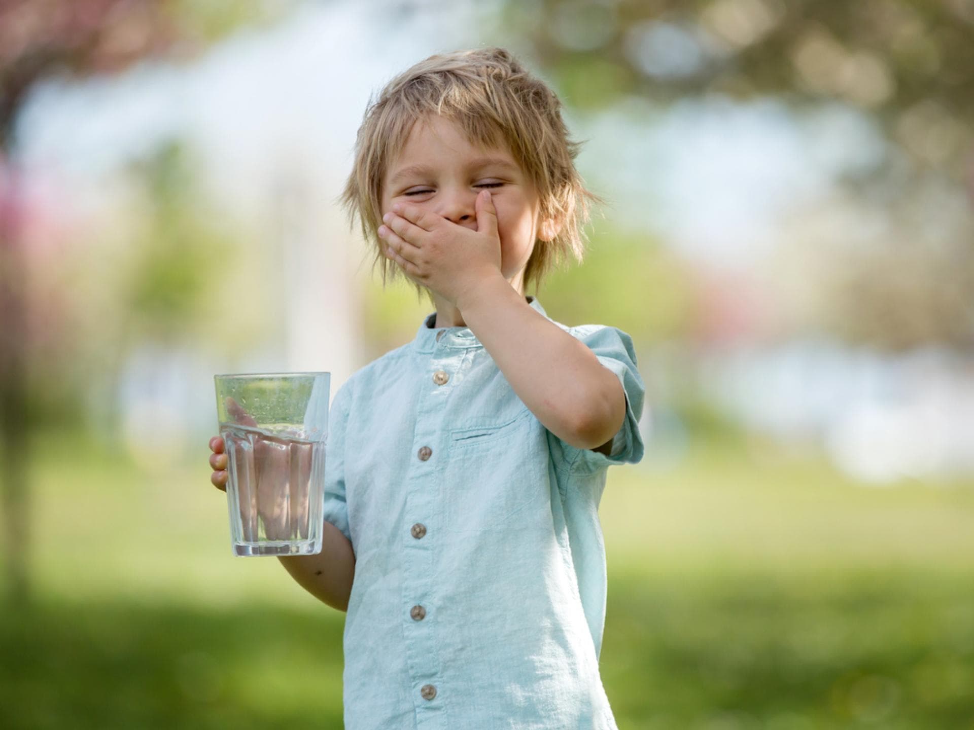 An mehreren Anzeichen merken Eltern, wenn Kinder zu wenig getrunken haben. Blonder Junge trinkt Wasser im Park.