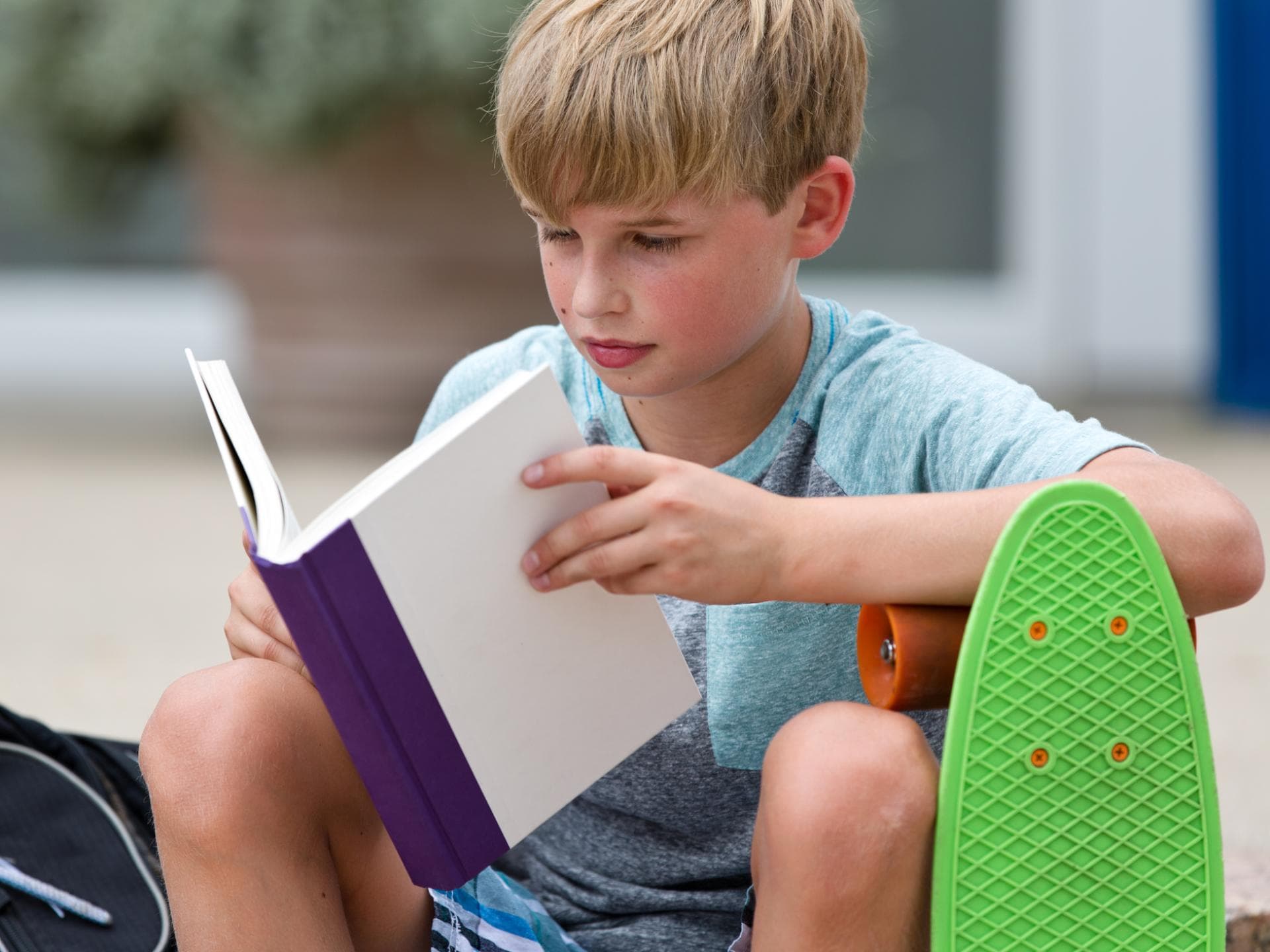 Junge mit Skateboard liest in einem Buch.