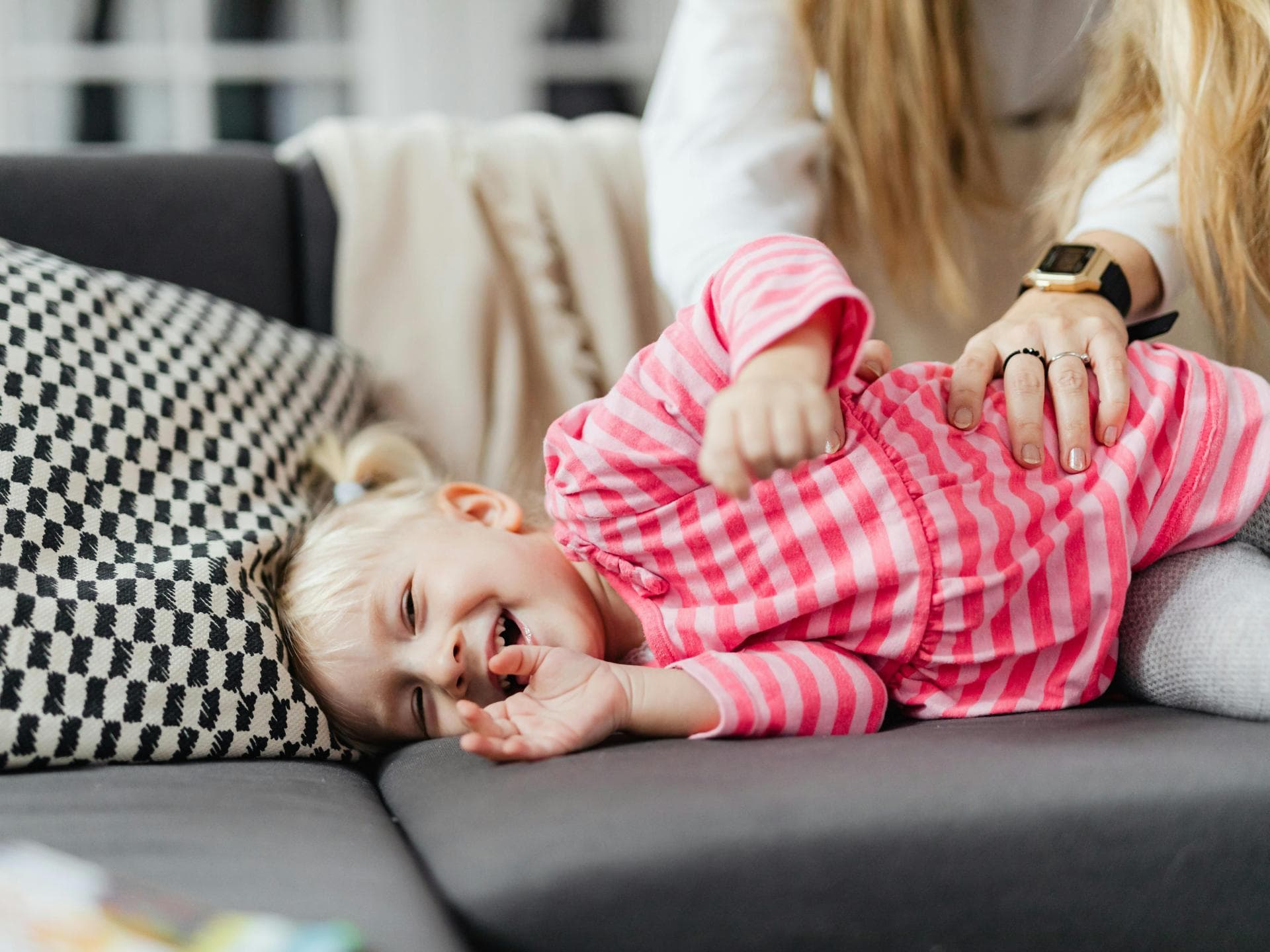 Ein kleines Mädchen wird von seiner Mutter auf dem Sofa gekitzelt.