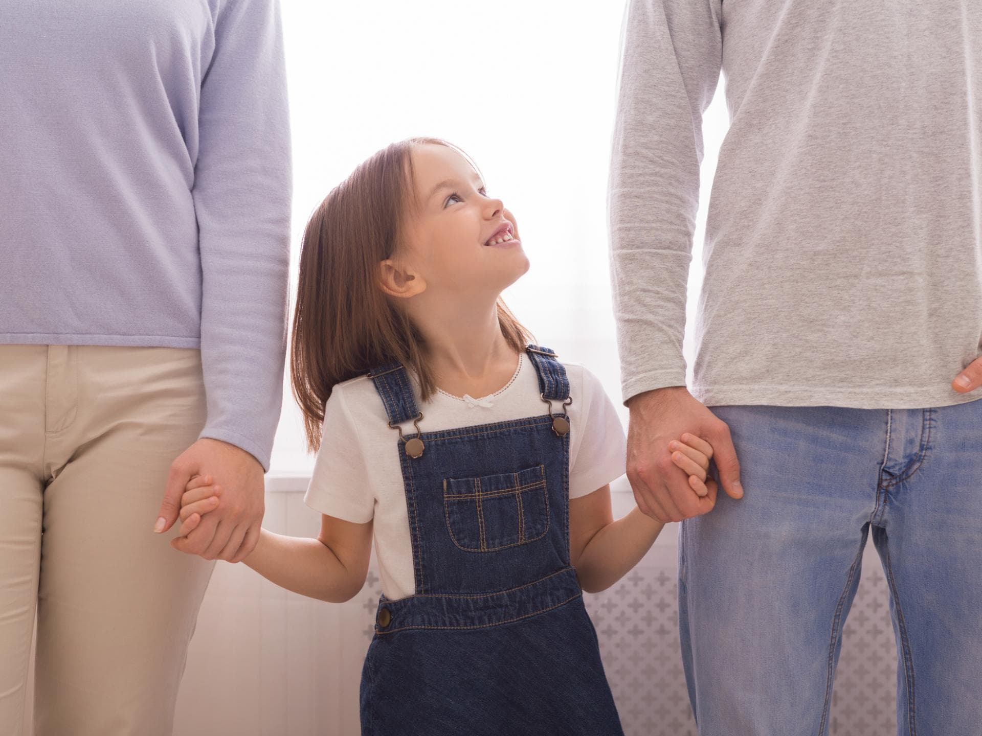 Das Aufwachsen als Einzelkind bringt besondere Herausforderungen mit sich. Lächelndes Mädchen steht zwischen ihren Eltern.