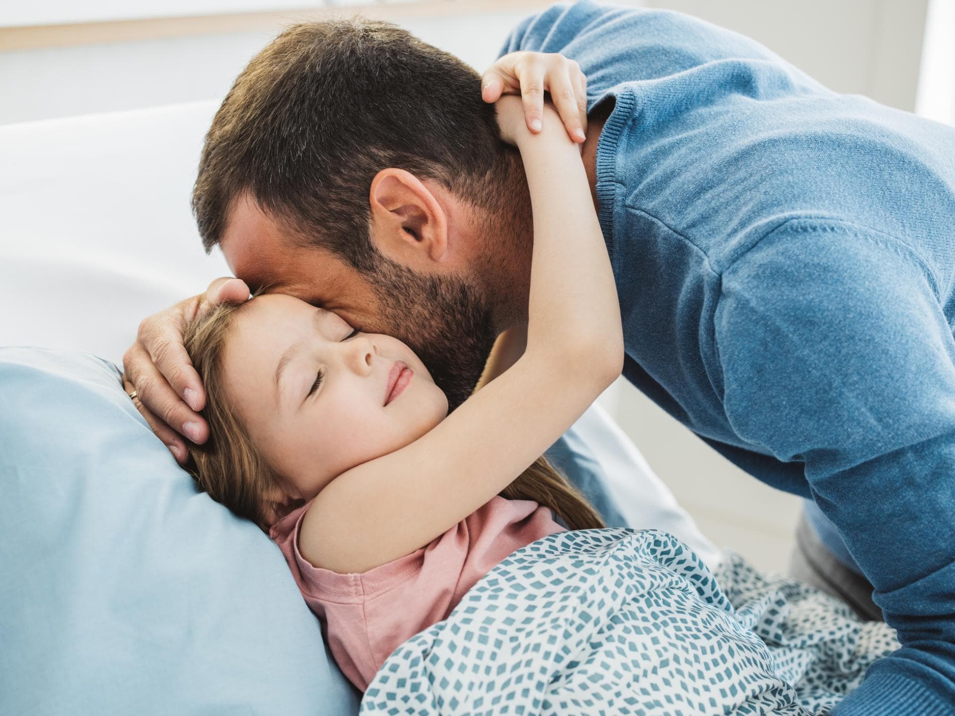 Liebevolles Kuscheln am Bett – das gefällt wohl allen Kindern. Kind umarmt seinen Vater beim Ins-Bett-Gehen.
