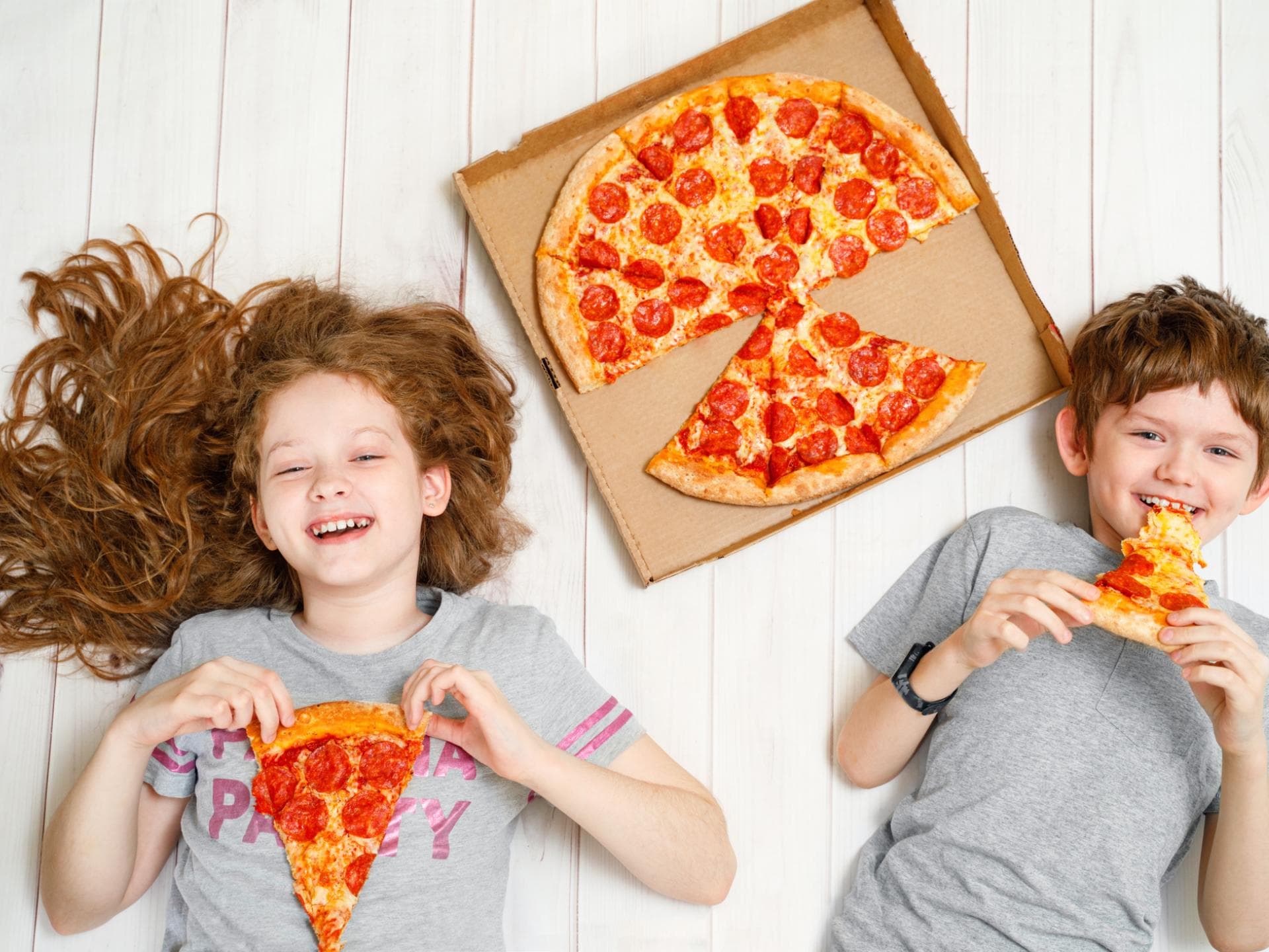 Da kommt Freude auf: Kinder lieben Salami-Pizza. Zwei fröhliche Kinder liegen mit Salami-Pizza auf dem Boden.
