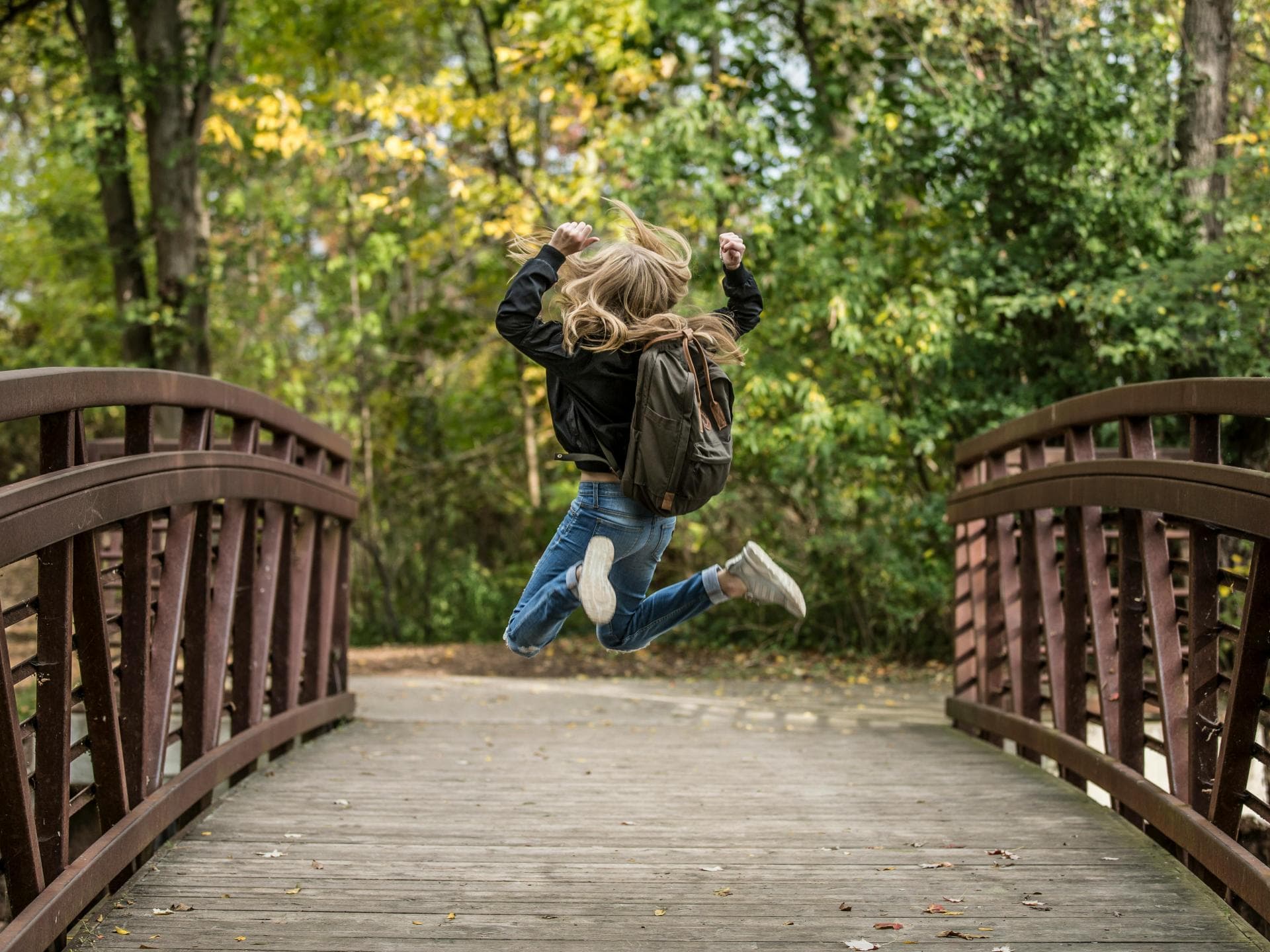 Mädchen mit Schulranzen springt hoch auf einer Holzbrücke.