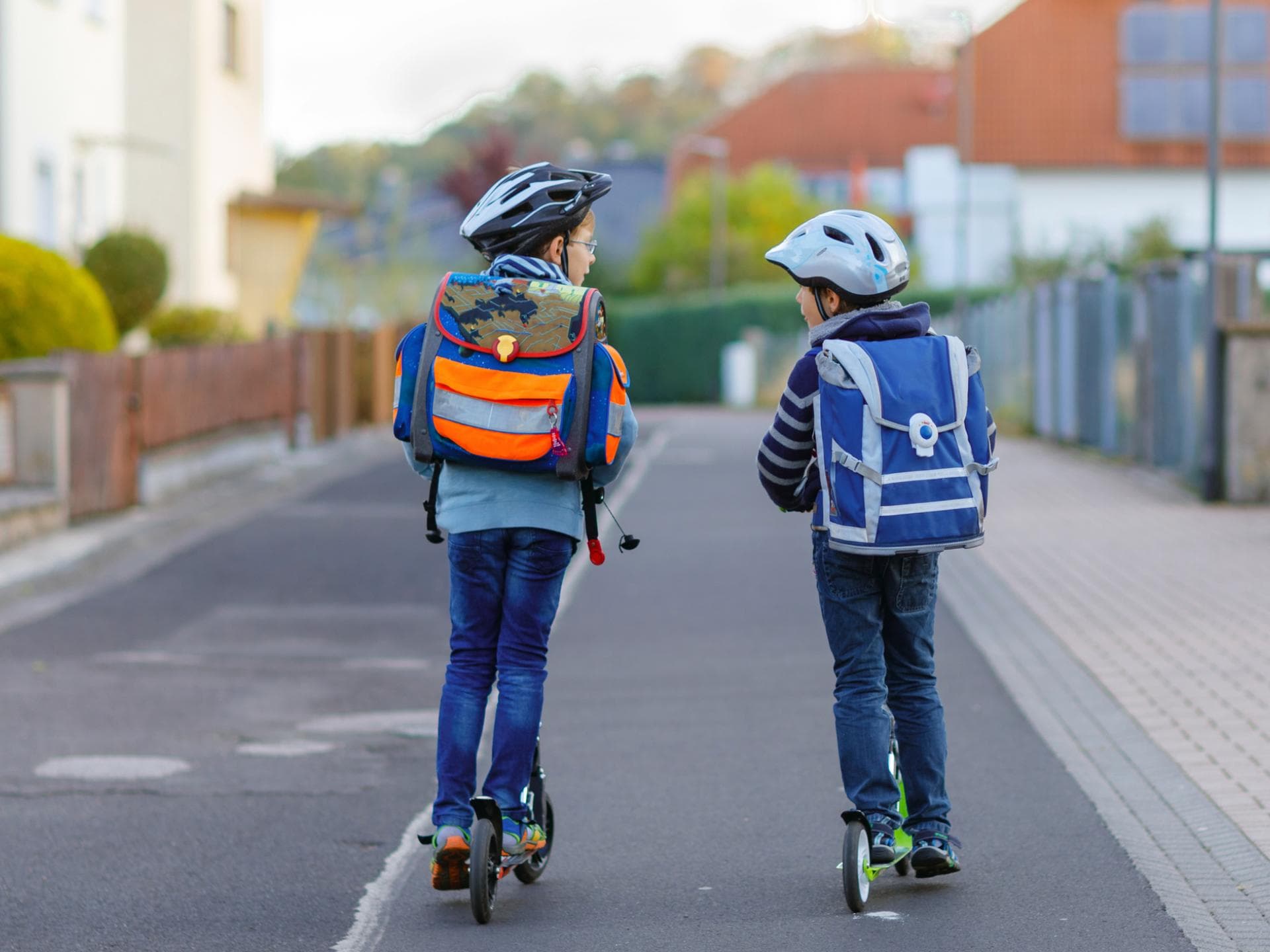 Zwei Kinder mit Schulranzen auf dem Rücken fahren auf Rollern zur Schule.