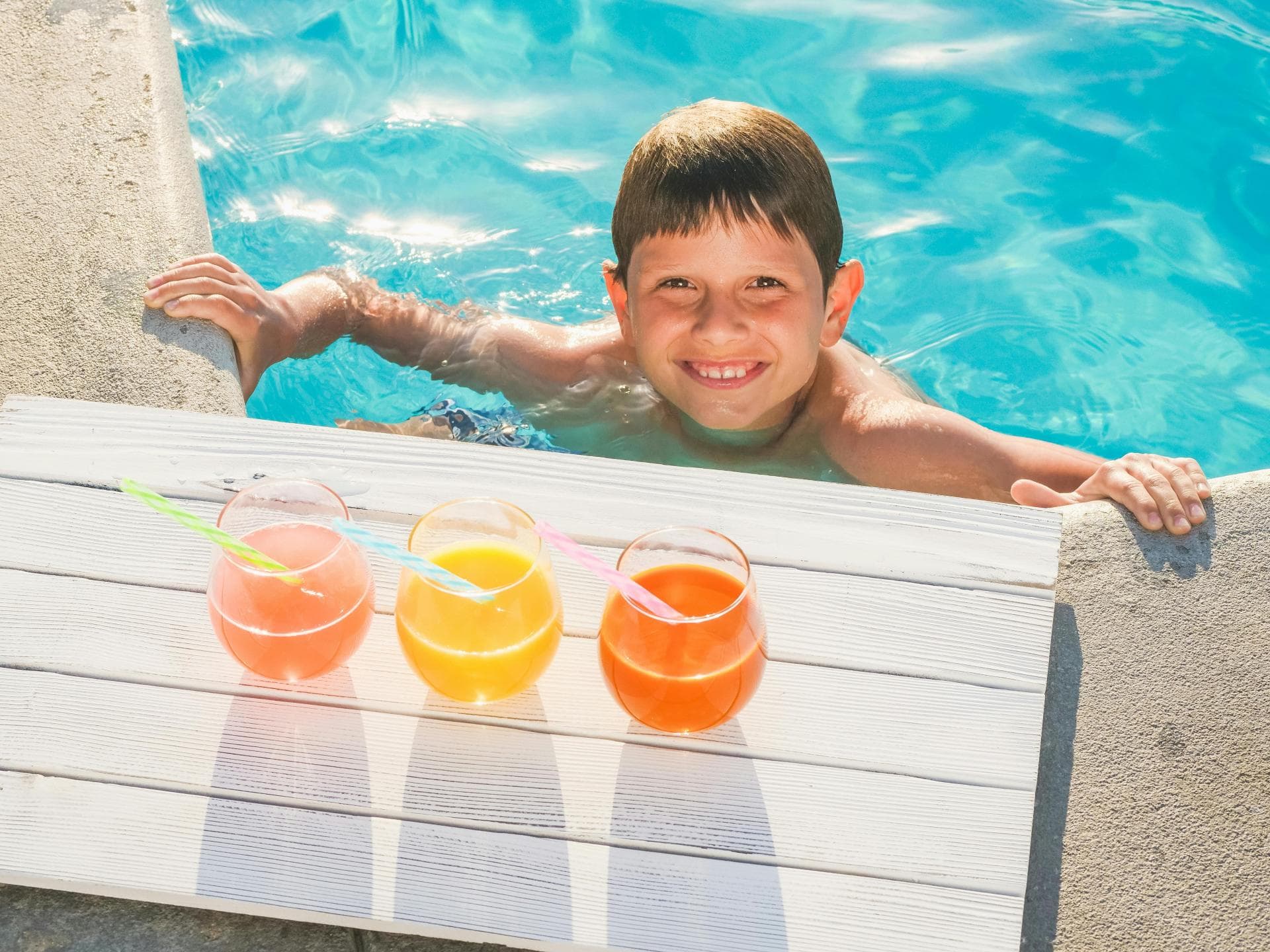 Junge im Pool vor drei Gläsern Saft.