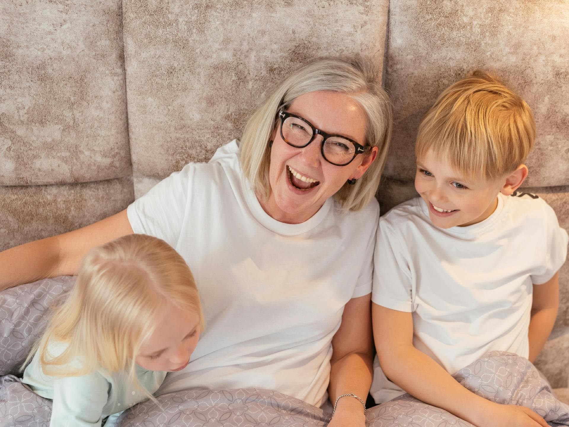 Eine Oma sitzt lachend mit ihren zwei Enkelkindern auf dem Sofa.