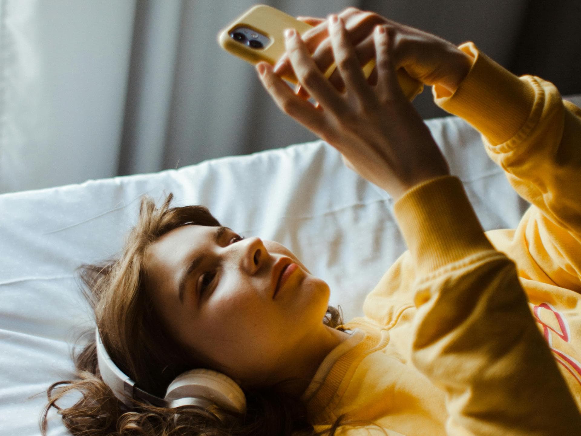Zu verlockend: Bildschirmzeit statt schlafen zu gehen. Frau im Bett am Handy mit Kopfhörern.