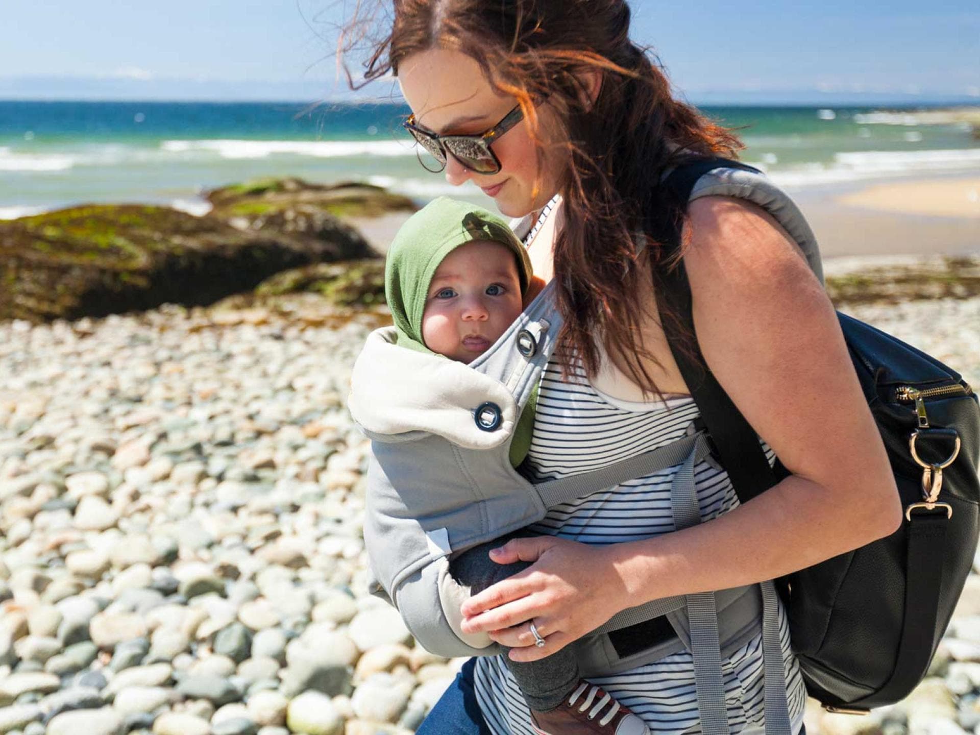 Im Sommer sollte euer Baby in der Trage weder schwitzen noch einen Sonnenbrand erleiden. Mutter mit Baby im Tragesitz am Strand