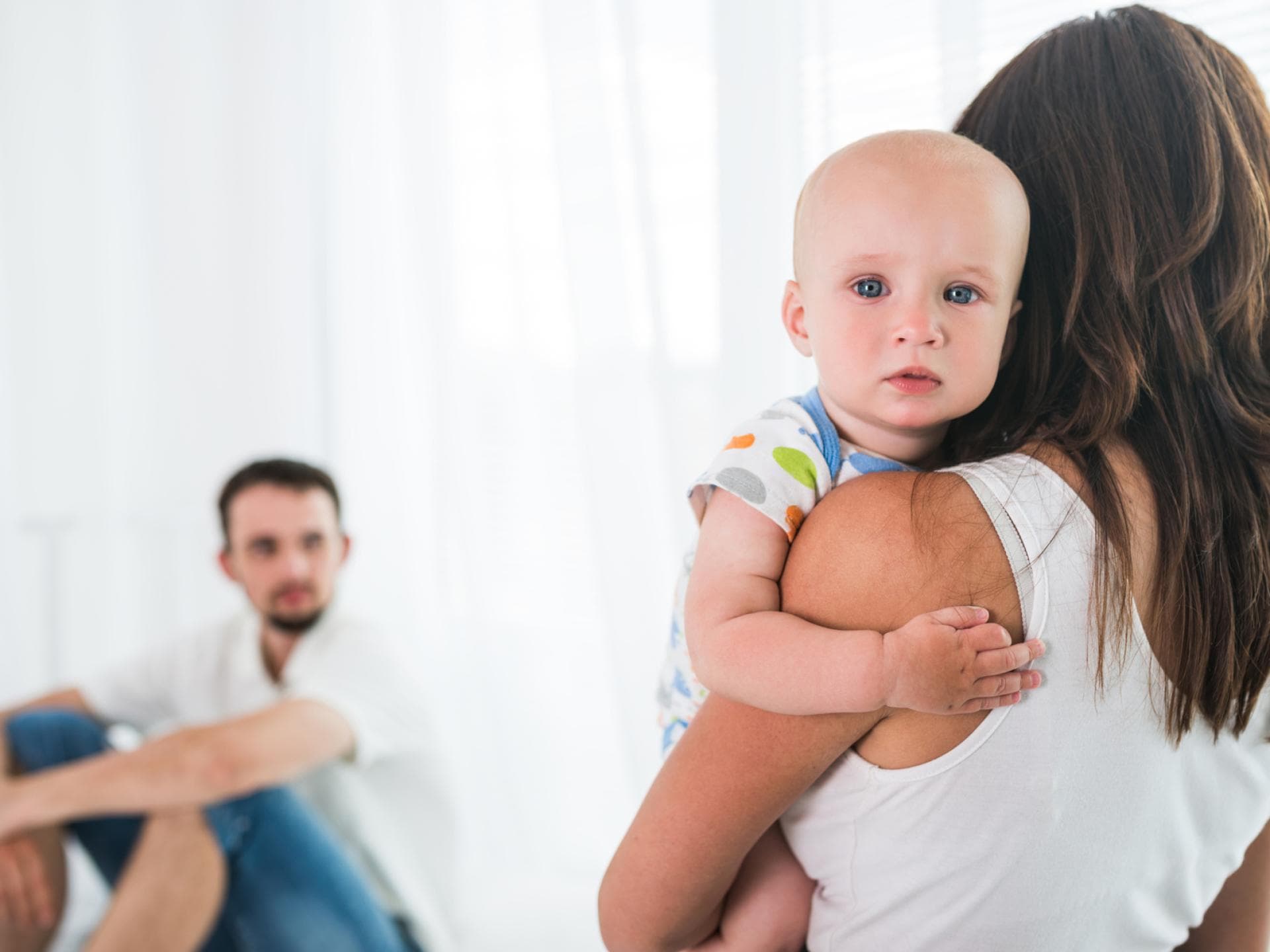 Mutter hält Baby auf dem Arm, Vater sitzt im Hintergrund.