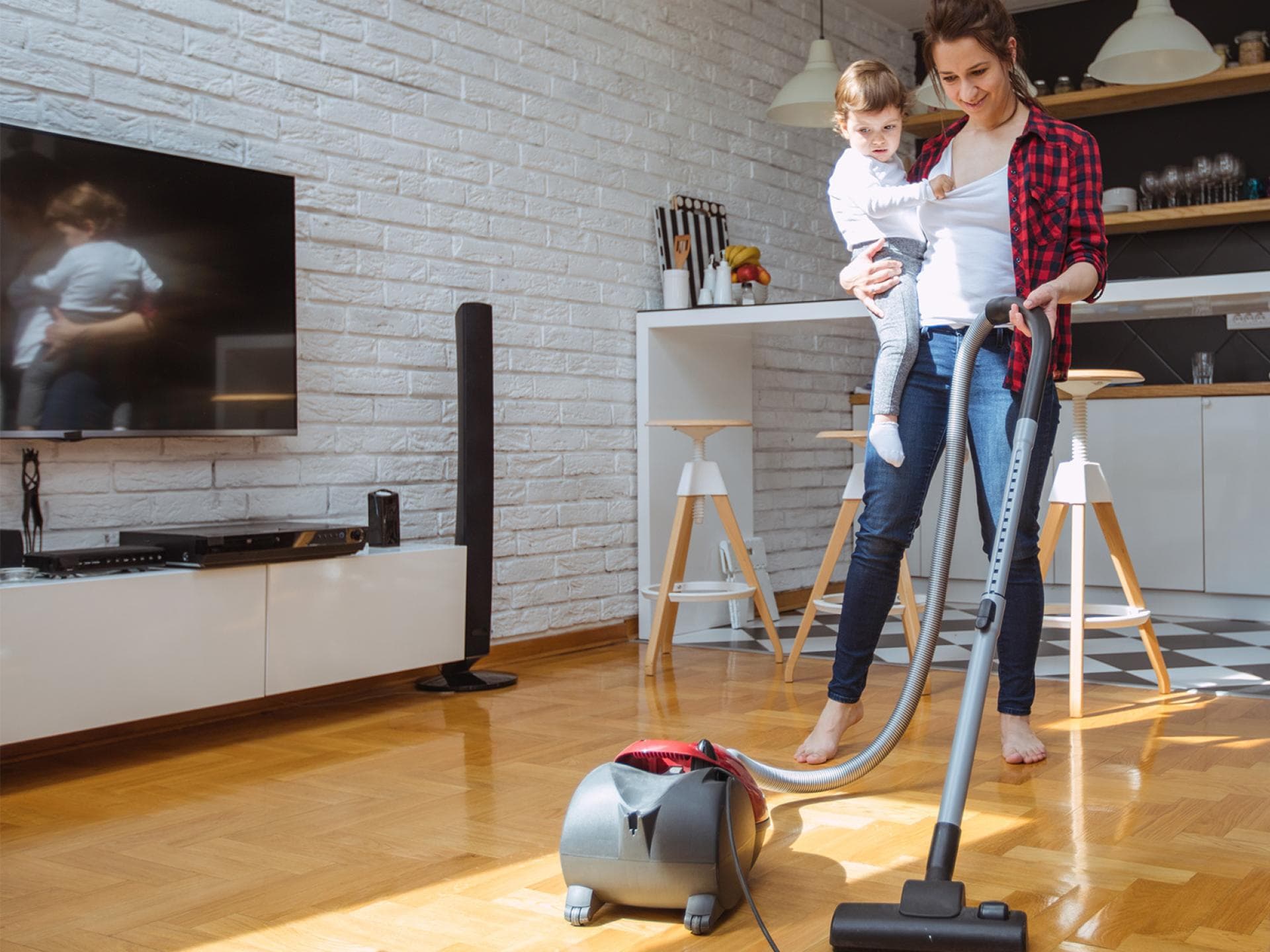 Multitasking – Mütter erledigen oft mehrere Jobs gleichzeitig. Gehalt gibt es dafür nicht. Mutter saugt mit Kleinkind auf dem Arm Staub.