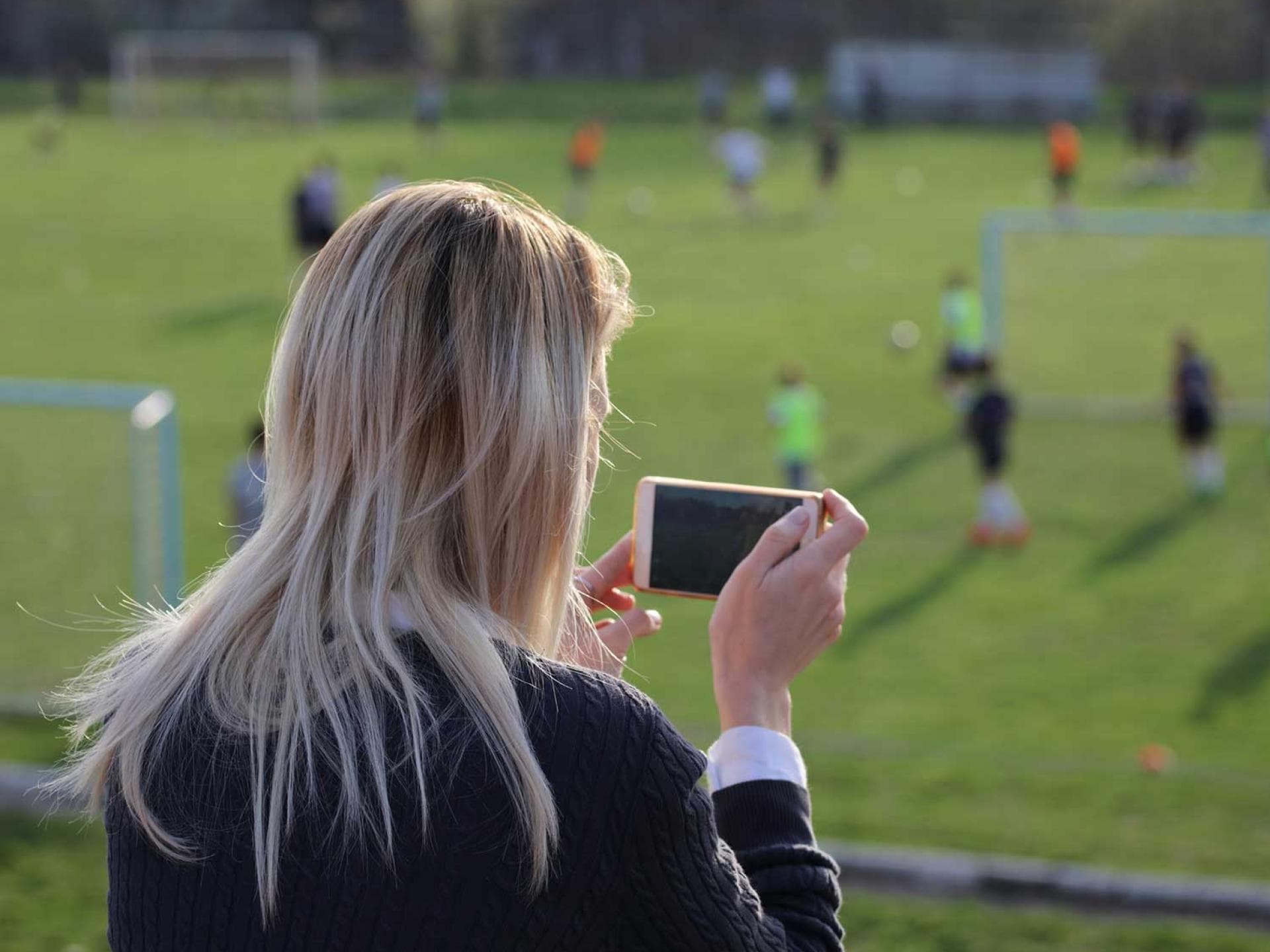 Ob Regen oder Sonnenschein: Mama ist dabei, wenn Sohnemann kickt. Mutter macht mit dem Smartphone Fotos vom Fußballspiel des Nachwuchses.