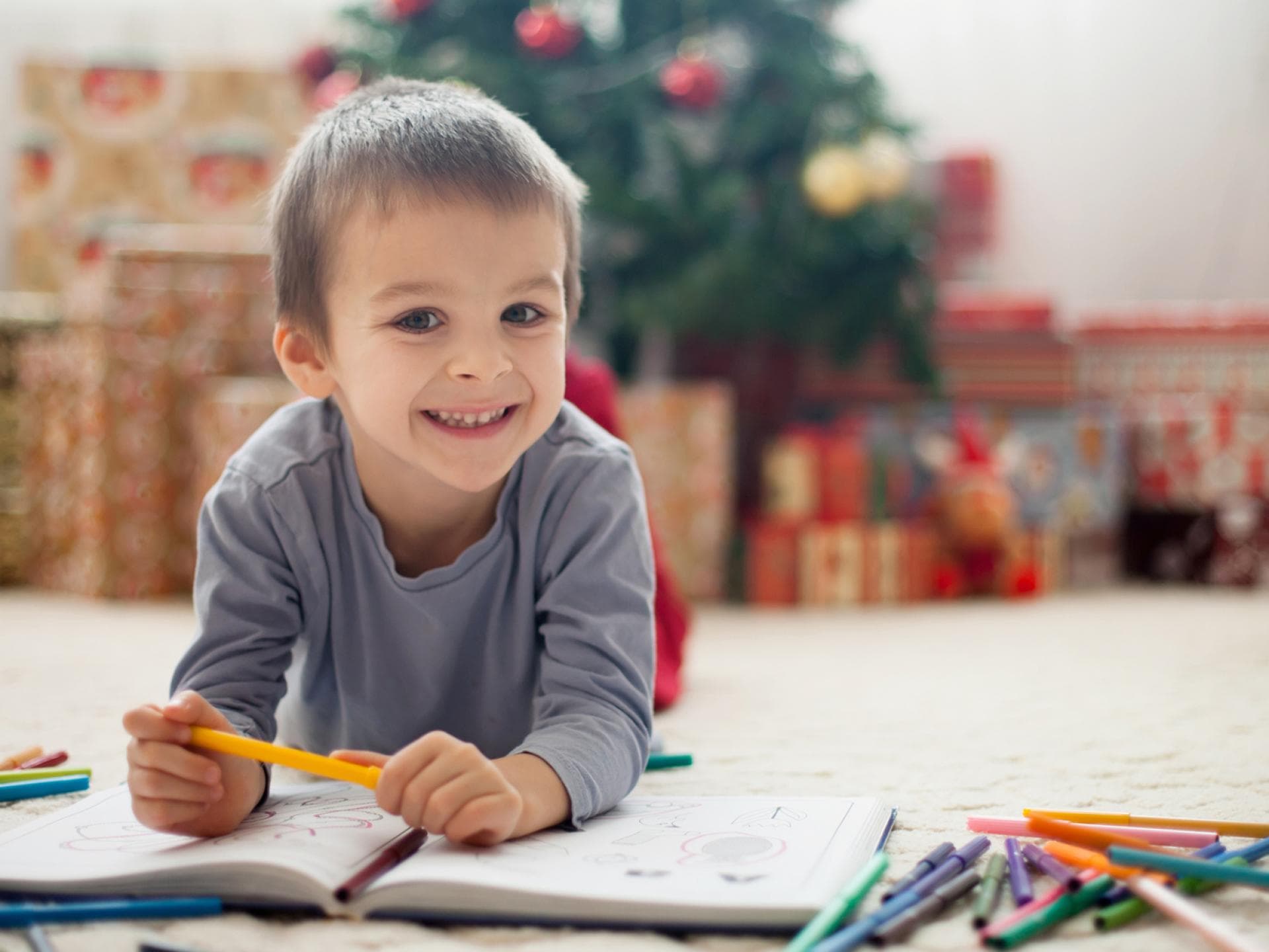 Weihnachtliche Ausmalbilder sind bei Kindern sehr beliebt. Ein kleiner Junge liegt vor dem Weihnachtsbaum auf dem Boden und malt Bilder..