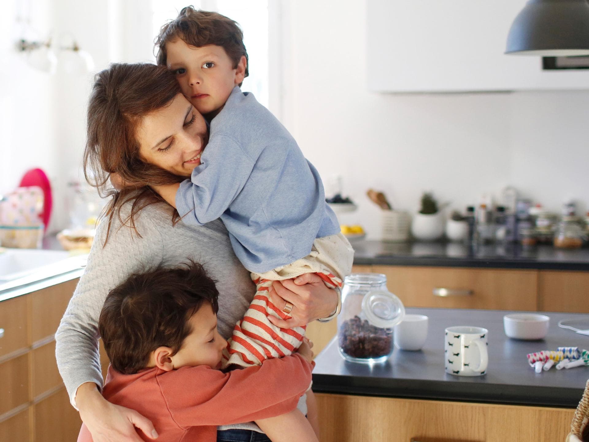 Gerade in unsicheren Zeiten brauchen Kinder viele Kuscheleinheiten. Mutter kuschelt in der Küche mit ihren beiden Kindern.